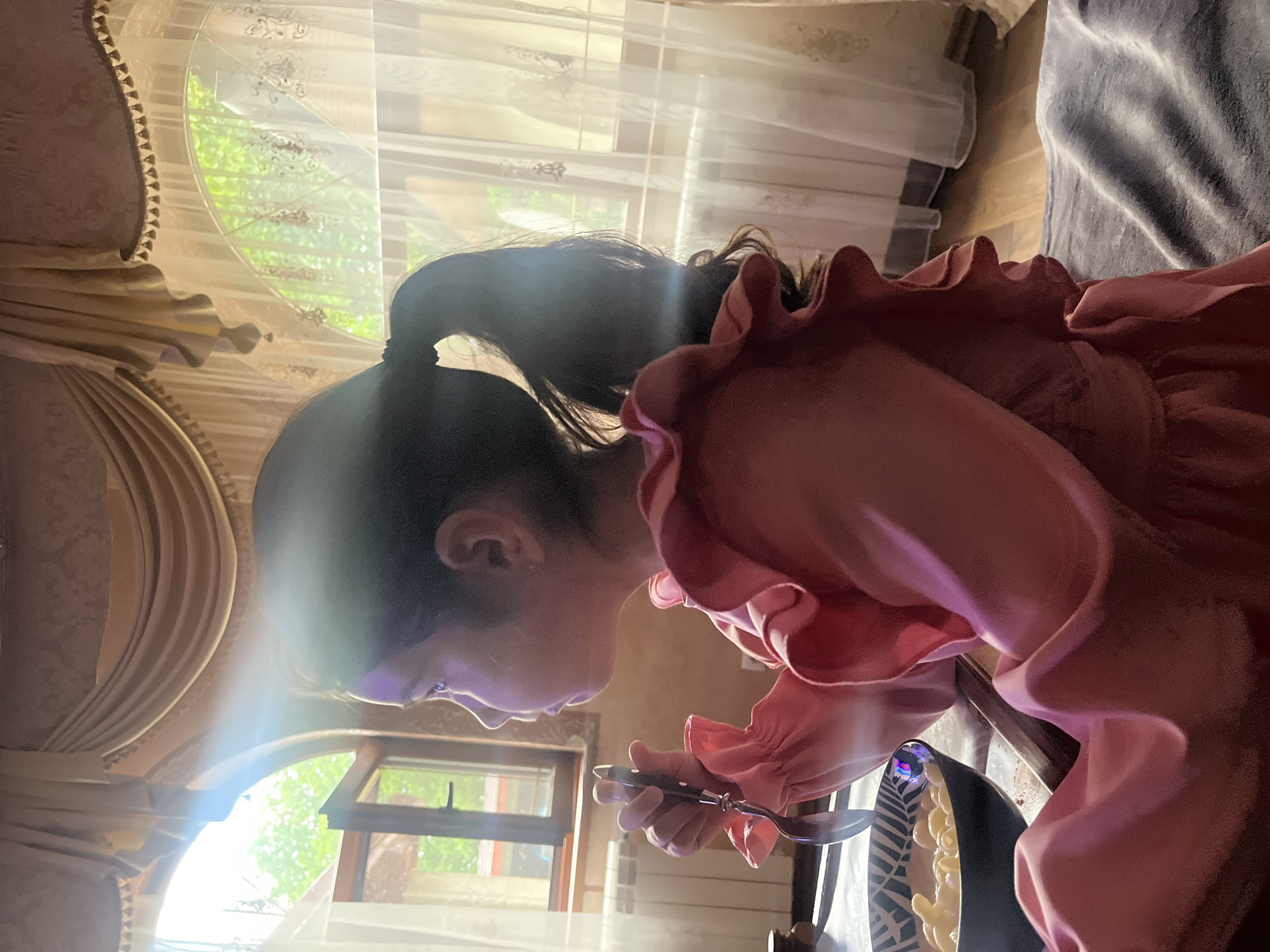 The image depicts a young girl in a pink ruffled dress seated at a table, holding a spoon and looking to her left, presumably at a window. She is facing the camera in profile. In front of her is a bowl of pasta. The background shows a window with curtains, suggesting an indoor setting. The light streaming through the window indicates it is daytime. The girl appears to be eating or about to eat. The scene is consistent with a domestic environment in Tarnivtsi, Ukraine, potentially a home or a restaurant with a view of the outdoors.