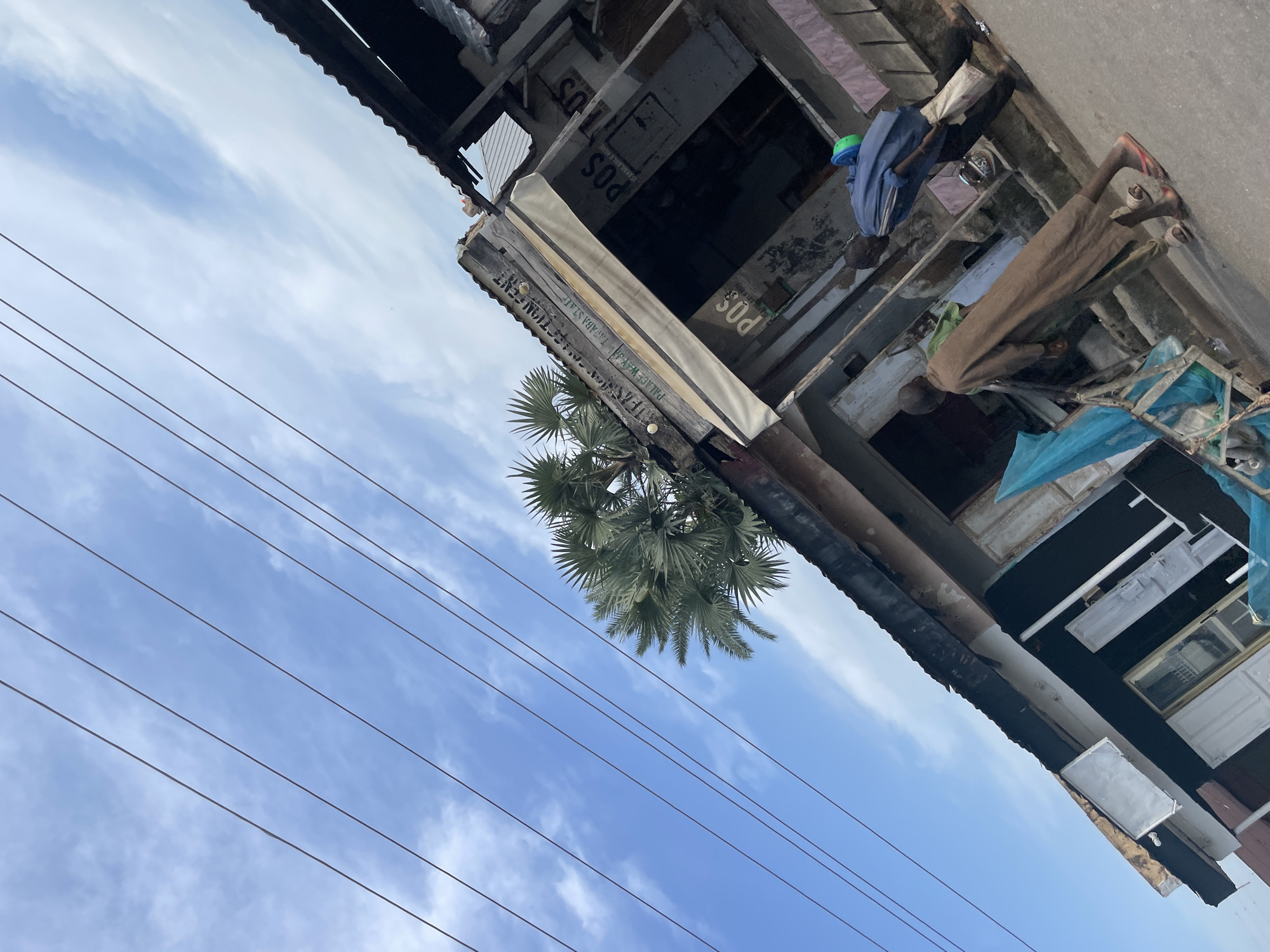 The image shows a street scene in Jalingo, Nigeria, viewed from a low angle, looking upwards. The sky is a bright blue with scattered white clouds, suggesting it is daytime with fair weather. Several power lines run diagonally across the frame.
The main focus is an outdoor structure, possibly a shop or stall, made of salvaged materials, with a corrugated metal roof. The structure appears weathered and somewhat dilapidated. Several signs are visible. One sign reads "PALACE WAY JA COTION CENT TAPABA STAT". Another sign has "POS" printed on it multiple times, with some text below it that is partially obscured but appears to say "IS AVAILABLE HERE" and "AMBLE MERE".
Two people are visible in the scene. One person is wearing a blue shirt and a green cap, and the other is wearing a brown garment. They appear to be standing or moving within the structure. The presence of people, the signs for "POS" (likely referring to Point of Sale services), and the general appearance of the setting suggest a commercial or community gathering spot in a semi-urban or rural area of Nigeria. The overall impression is one of everyday life in a bustling, somewhat rustic environment.