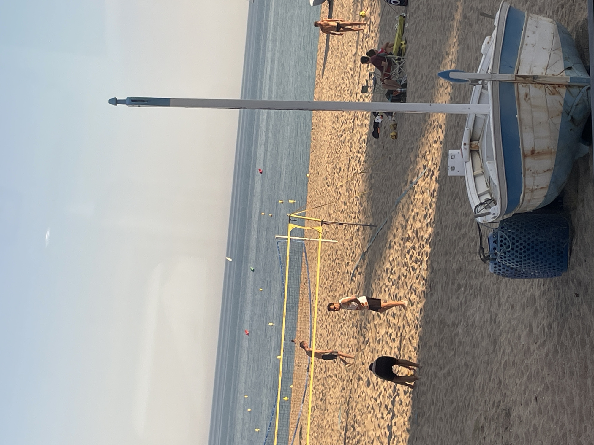 The image depicts a beach scene in Sant Pol De Mar, Spain. A small, white and blue boat rests on the sandy beach near a blue plastic basket. A volleyball net is visible, with people playing or walking nearby. In the background, the sea stretches out to the horizon under a clear sky. Several individuals are seen sitting or walking along the beach.