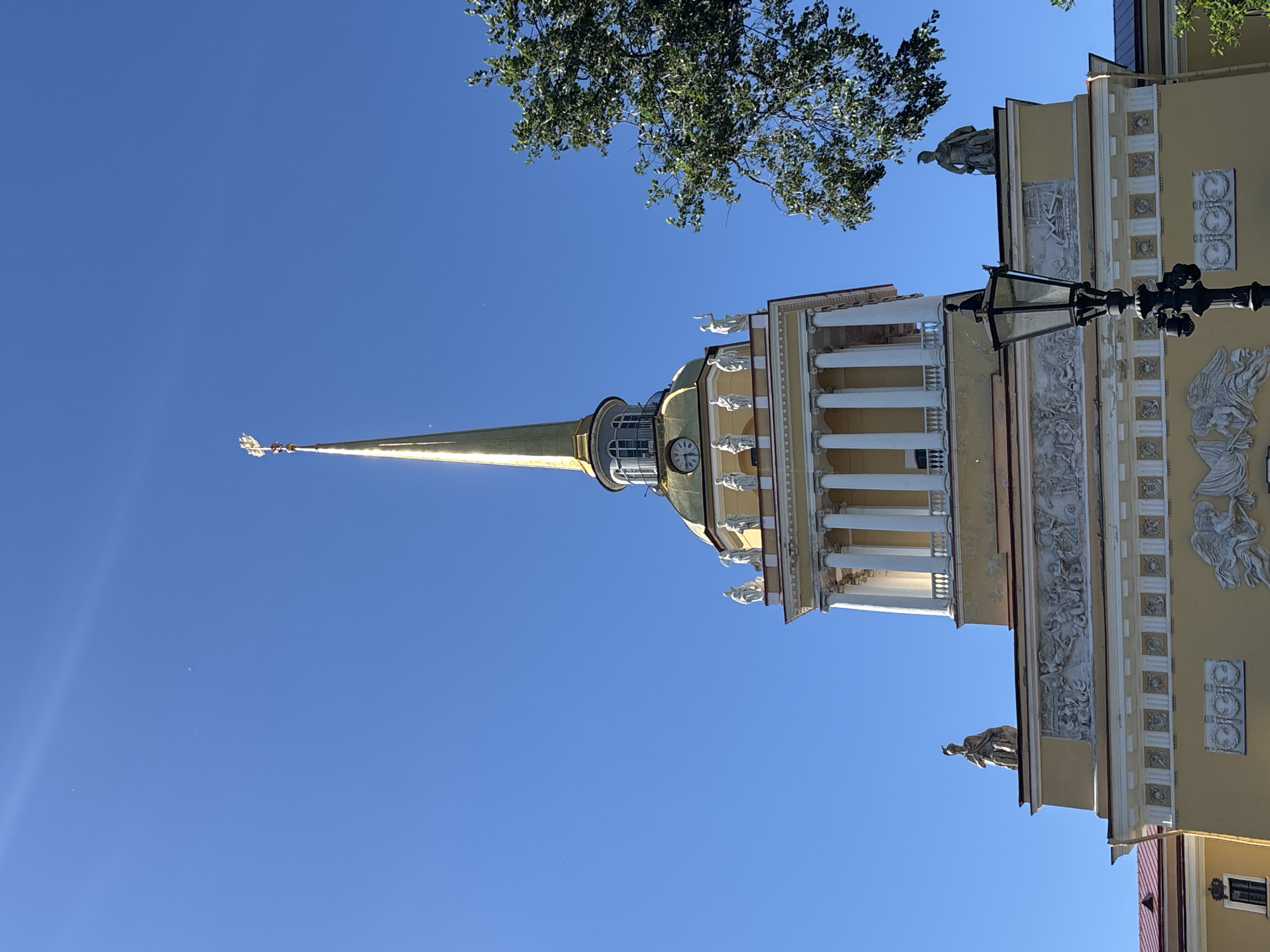 This is a low-angle, rotated shot of the Admiralty Building in Saint Petersburg, Russia. The main subject is the iconic golden spire of the building, which dominates the frame against a clear, bright blue sky. The spire is topped with a weather vane, and a small flag is visible near its base.
The building itself, a pale yellow color, features classical architecture with white columns, decorative friezes depicting mythological scenes, and numerous statues of winged figures and seated individuals adorning its facade. A clock is visible on a rounded section of the building near the base of the spire. A black, ornate lamppost is positioned in the foreground, adding a touch of architectural detail. In the upper left corner, a portion of a leafy green tree is visible, suggesting a pleasant, possibly warm day. The bright sunlight and clear sky indicate it is daytime, likely late morning or early afternoon. The overall impression is one of grandeur and historical significance. No visible text is present in the image.