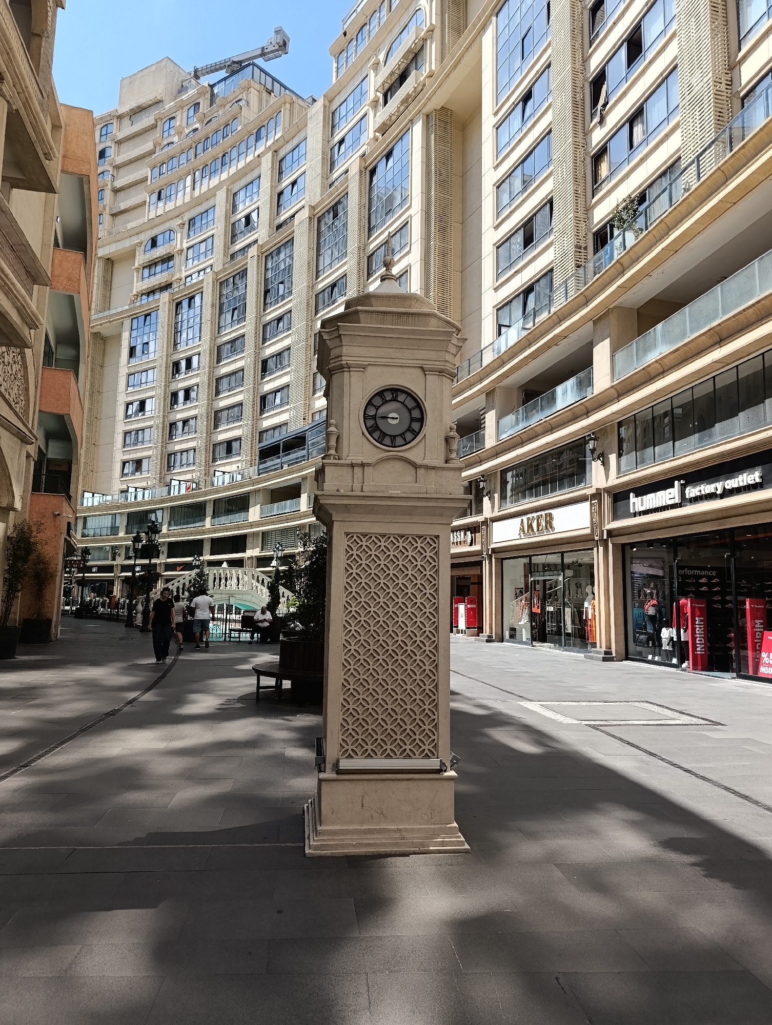 In Gaziosmanpaşa, Turkey, a pedestrian walkway is lined with modern buildings and shops on a sunny day. A prominent stone clock tower stands in the foreground on the right, featuring an ornate patterned base and a clock face with Roman numerals. To the left, a narrow alleyway leads to a wider street where several people are walking. In the background, tall, multi-story buildings with numerous windows dominate the skyline. Shops with illuminated signs like "AKER" and "hummel factory outlet" are visible on the ground floor. A white pedestrian bridge crosses further down the street, and several benches are placed along the walkway. The pavement is paved with large, grey tiles, and shadows from trees and buildings are cast across the scene. A construction crane is visible atop one of the buildings in the distance.
