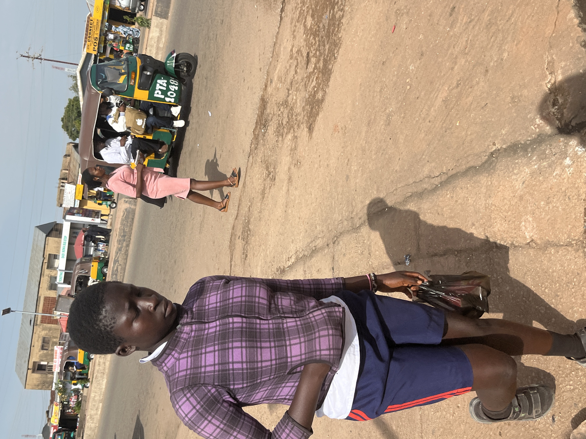 In Gura Top, Nigeria, a young person stands on a dusty, sun-drenched road. They wear a purple and black plaid zip-up jacket, a white undershirt, and navy blue shorts with red stripes down the sides. Their hands are in the pockets of the jacket, and they hold a dark plastic bag in their right hand. They are wearing dark sandals and black socks. In the background, a woman in a pink dress walks past a bright green and yellow auto-rickshaw. The auto-rickshaw is marked with "PTA 1048" and appears to be carrying passengers. Buildings with corrugated metal roofs line the street, some displaying signs for businesses like "POS" and "CONNECT." The scene captures a typical day in a busy urban environment, with pedestrians and vehicles navigating the roadways.