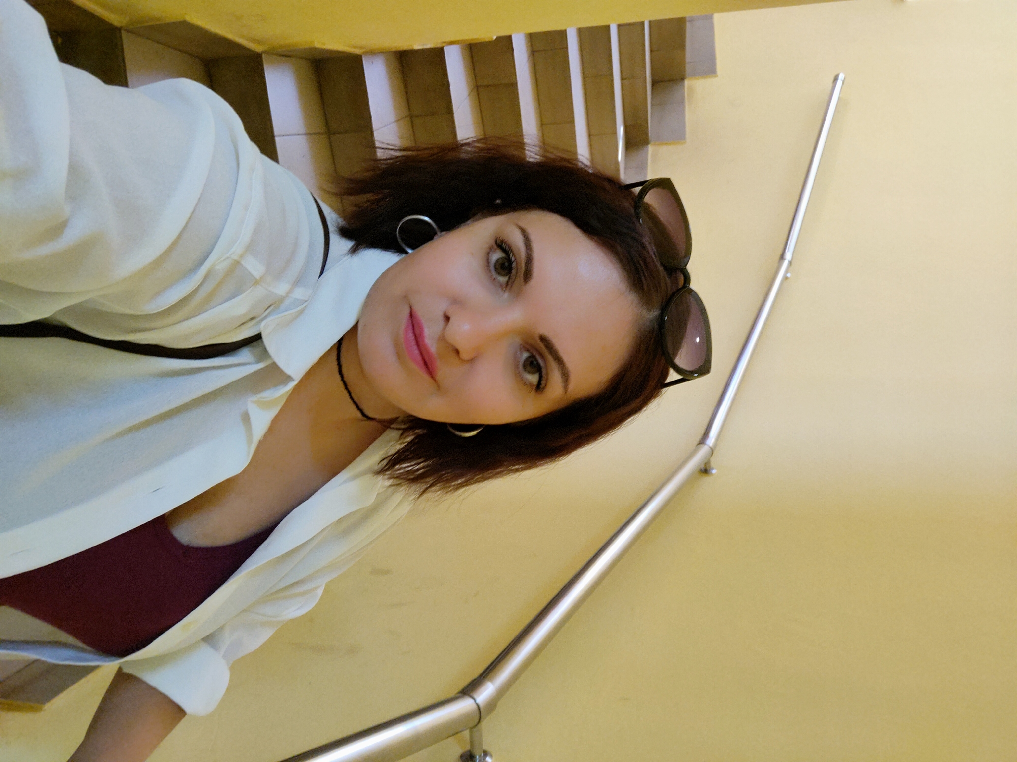 A woman with short dark hair, wearing a white shirt over a maroon top and sunglasses on her head, is taking a selfie on a staircase. The staircase has light yellow walls and metal railings. The location is Chaikovsky, Russia.