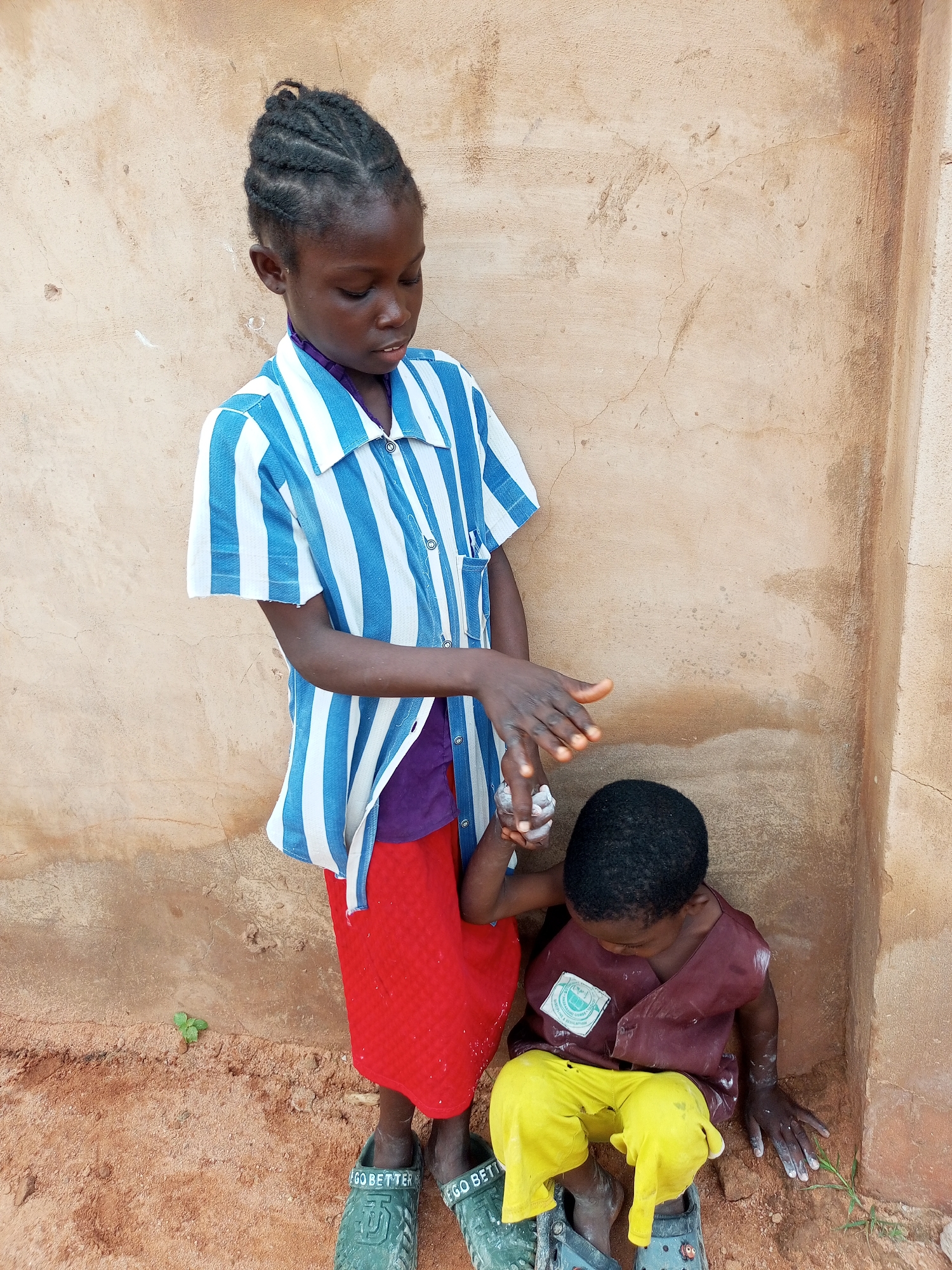 In Tempure, Nigeria, a young Black girl with braided hair stands to the left of the frame, wearing a blue and white striped short-sleeved button-down shirt over a purple shirt and a red skirt. Her right hand is extended downwards, interacting with a younger Black child who is seated on the ground to her right. This younger child wears a maroon sleeveless vest over bright yellow pants. The child's left hand is extended outwards, touching a small green plant growing in the sandy soil. Both children are wearing green open-toed shoes. The background is a textured, light brown wall with some visible cracks and darker, stained areas. The ground is composed of reddish-brown soil with scattered small stones and dirt. The scene appears to be outdoors, in a somewhat rural or less developed urban environment.