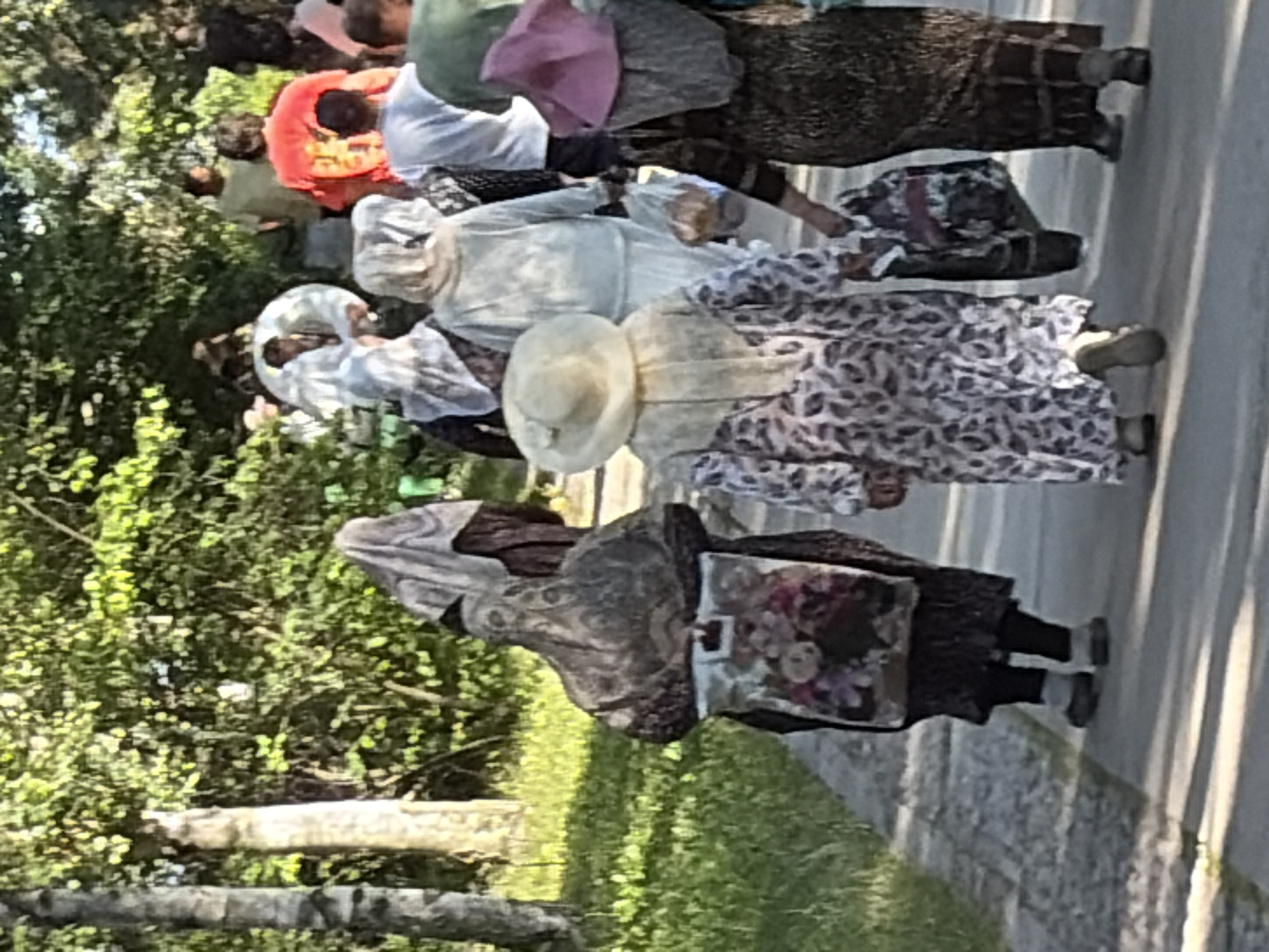 An outdoor daytime, eye-level shot captures a group of people, predominantly women, walking on a paved path bordered by grass and trees. The individuals are seen from behind, and most are wearing traditional head coverings and long dresses. Some carry bags. The lighting suggests it is a sunny day, with shadows cast on the path. The scene is set in a park-like environment, possibly within a city, though the specific urban location is unknown.
