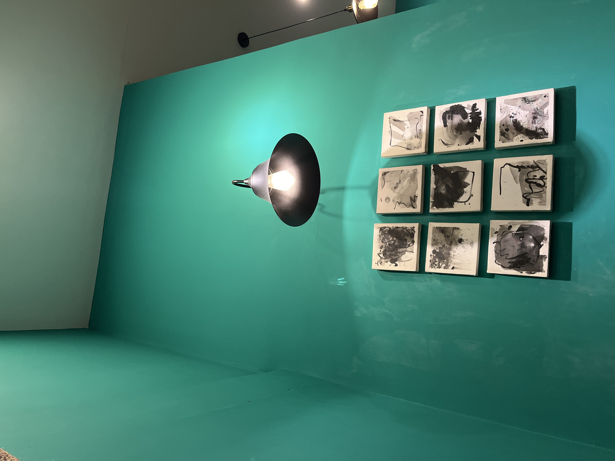 The image depicts an interior space with a teal-colored wall and floor. On the right wall, nine small, square artworks are arranged in a 3x3 grid. The artworks appear to be black and white abstract paintings. A black, conical lamp with a visible bulb is mounted on the wall to the left of the artworks. The wall in the background is painted a light green color. The location is not discernible from the image. There are no people visible.