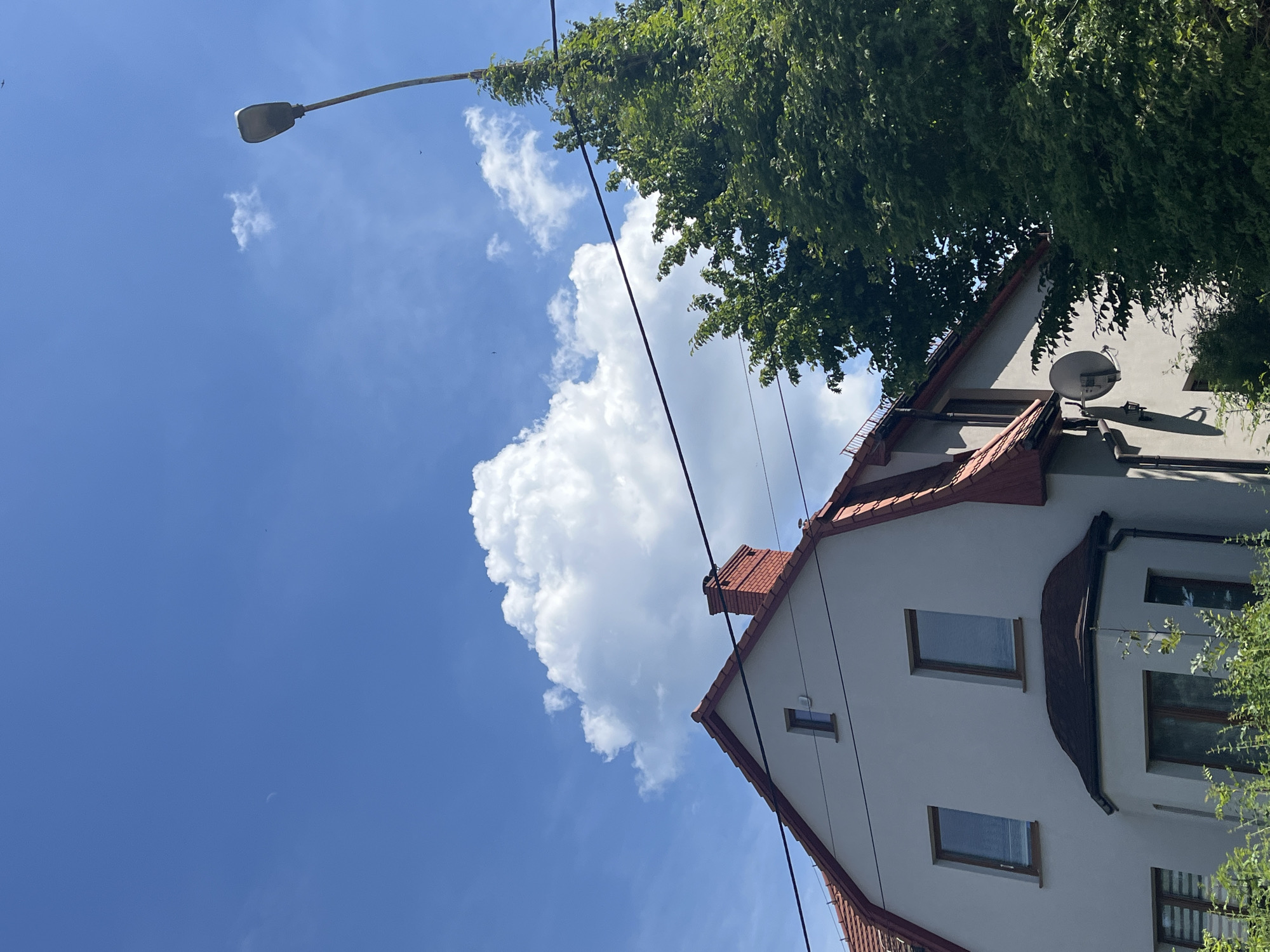This is a close-up, slightly tilted, outdoor shot of a white building with a red-tiled roof, set against a bright blue sky with fluffy white clouds. A large tree with dense green foliage partially obscures the upper portion of the building. A street lamp with a modern design is visible in the upper left quadrant, with a black cable running from it towards the building. The building features several windows, some with blinds visible. A satellite dish is mounted on the wall of the building. The scene appears to be set during the daytime, with clear weather. The overall impression is one of a residential area on a sunny day.