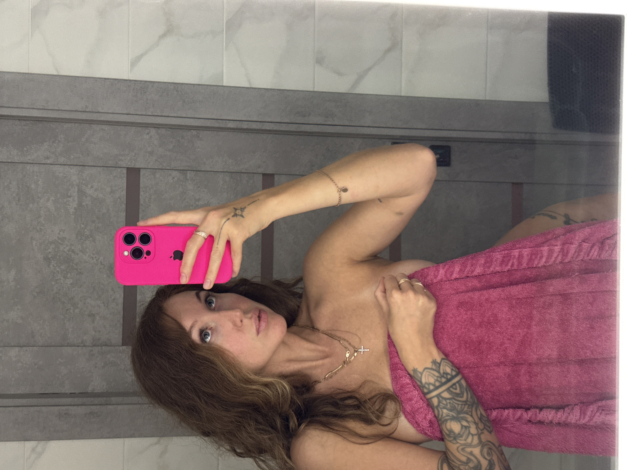 A woman with light skin and brown wavy hair is taking a selfie in a bathroom. She is holding a pink iPhone in her left hand, which is held up and slightly angled towards her face. Her left arm has a tattoo of a starburst on the wrist and a thin gold bracelet higher up. She has multiple rings on her fingers. The woman is wearing a gold necklace with a cross pendant. She is partially wrapped in a pink towel. Tattoos are visible on her right arm.
The background appears to be a bathroom with gray tiled walls. A mirror frame is partially visible along the edge of the scene.
The image provides no information about the city of Chaikovsky, Russia.
