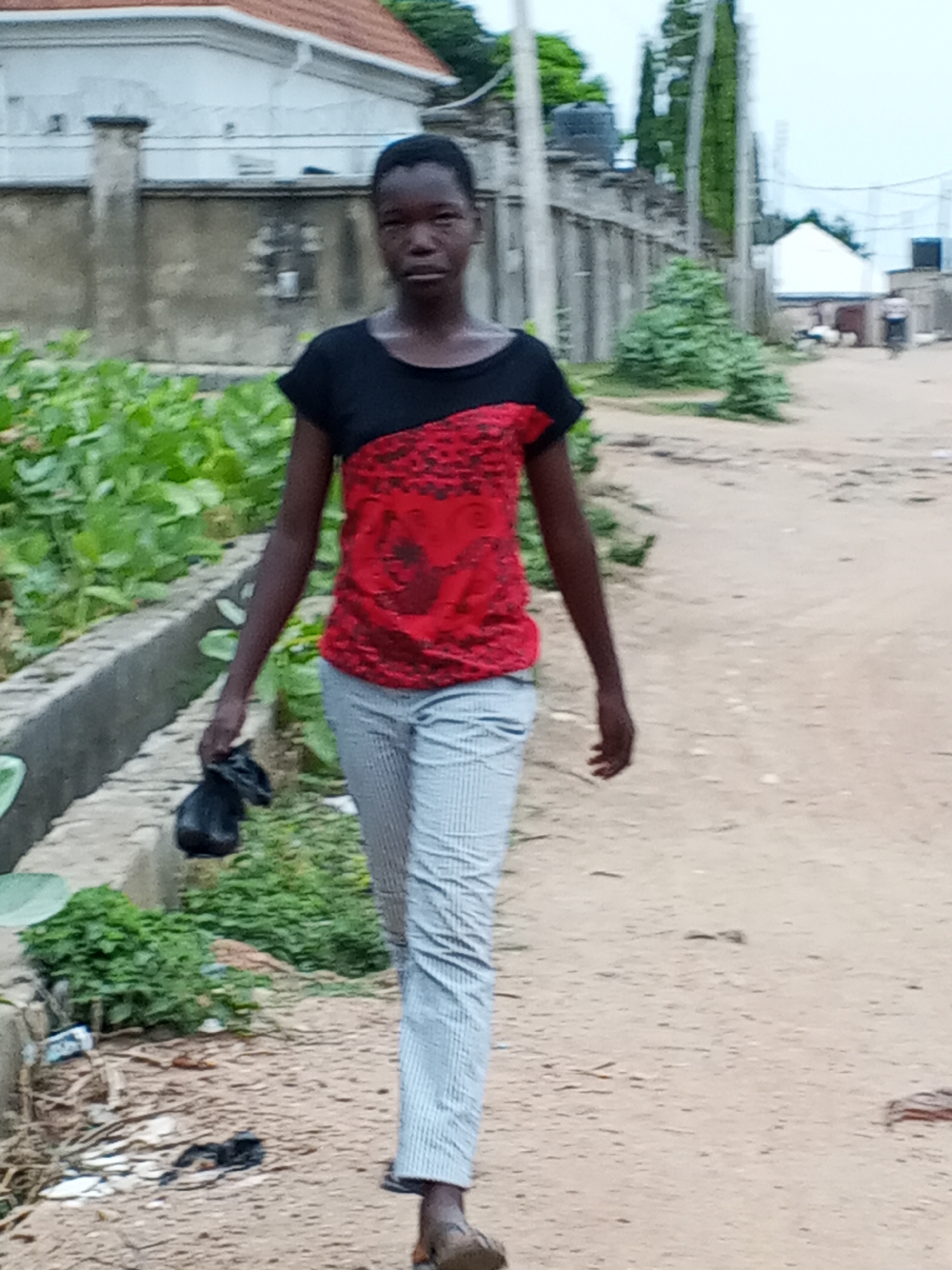 A young person walks down a dusty road in Otokiti, Nigeria. The individual is wearing a black and red shirt, striped pants, and sandals, and is carrying a small black bag in their left hand. Behind the person is a building with a red roof, a gray fence, and lush green plants along the side of the road. In the background, further down the road, are other buildings and a person on a bicycle.