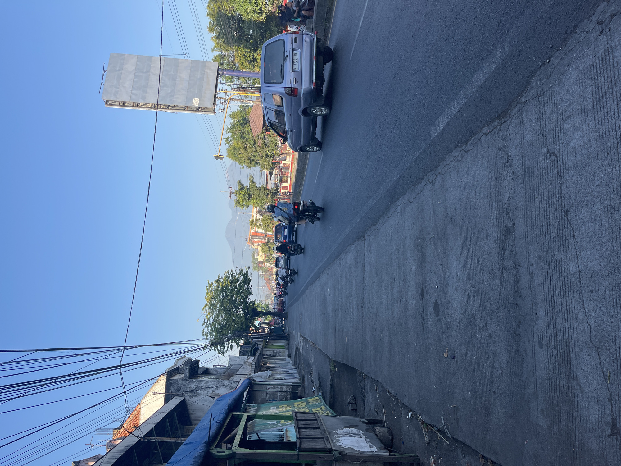 The image shows a street scene in Porong, Indonesia. On the left side of the frame, a sidewalk runs alongside a row of buildings with awnings and signage. The buildings appear to be shops or small businesses. The sidewalk is made of concrete and shows signs of wear and tear, with some debris scattered on it.
On the right side of the frame is a paved road with traffic. Several motorcycles and a silver SUV are visible, all moving in the same direction. The SUV has a license plate that reads "W 1733 RA". A traffic light and a large, blank billboard are also present on the right side of the road.
In the background, there are trees, more buildings, and in the distance, a mountain range is visible under a clear blue sky. The overall impression is of a bustling, everyday street in a tropical or subtropical urban environment. The lighting suggests it is either early morning or late afternoon, with long shadows cast on the sidewalk.