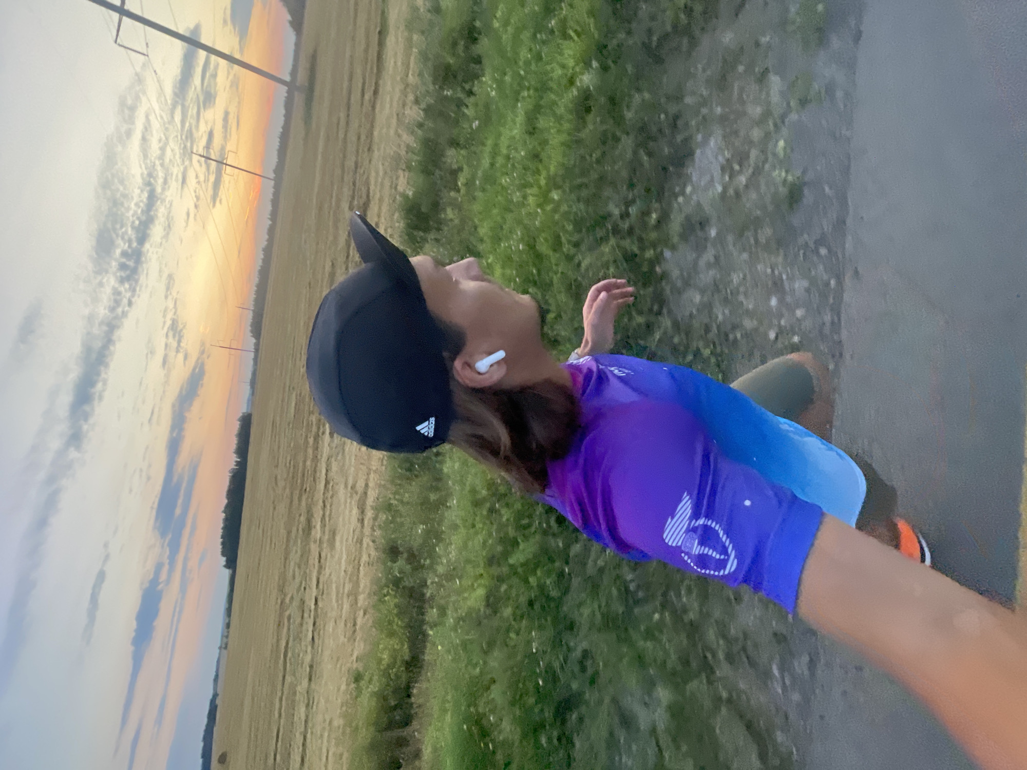 The image shows a person running outdoors during what appears to be sunset. The person is wearing a black baseball cap with an Adidas logo, wireless earbuds, a purple and blue gradient t-shirt with a white circular logo on the sleeve, and green shorts. They are also wearing orange and white athletic shoes. The person is captured in motion, suggesting they are mid-run.
The background features a harvested field, green vegetation lining a path or road, and a dramatic sky with clouds tinged with orange and pink from the setting sun. Tall utility poles with power lines are visible against the sky, indicating an outdoor, possibly rural or semi-rural, setting. The image is taken from a low angle, possibly by the runner themselves using a selfie perspective.
The location is identified as Ol'khovo, Russia.