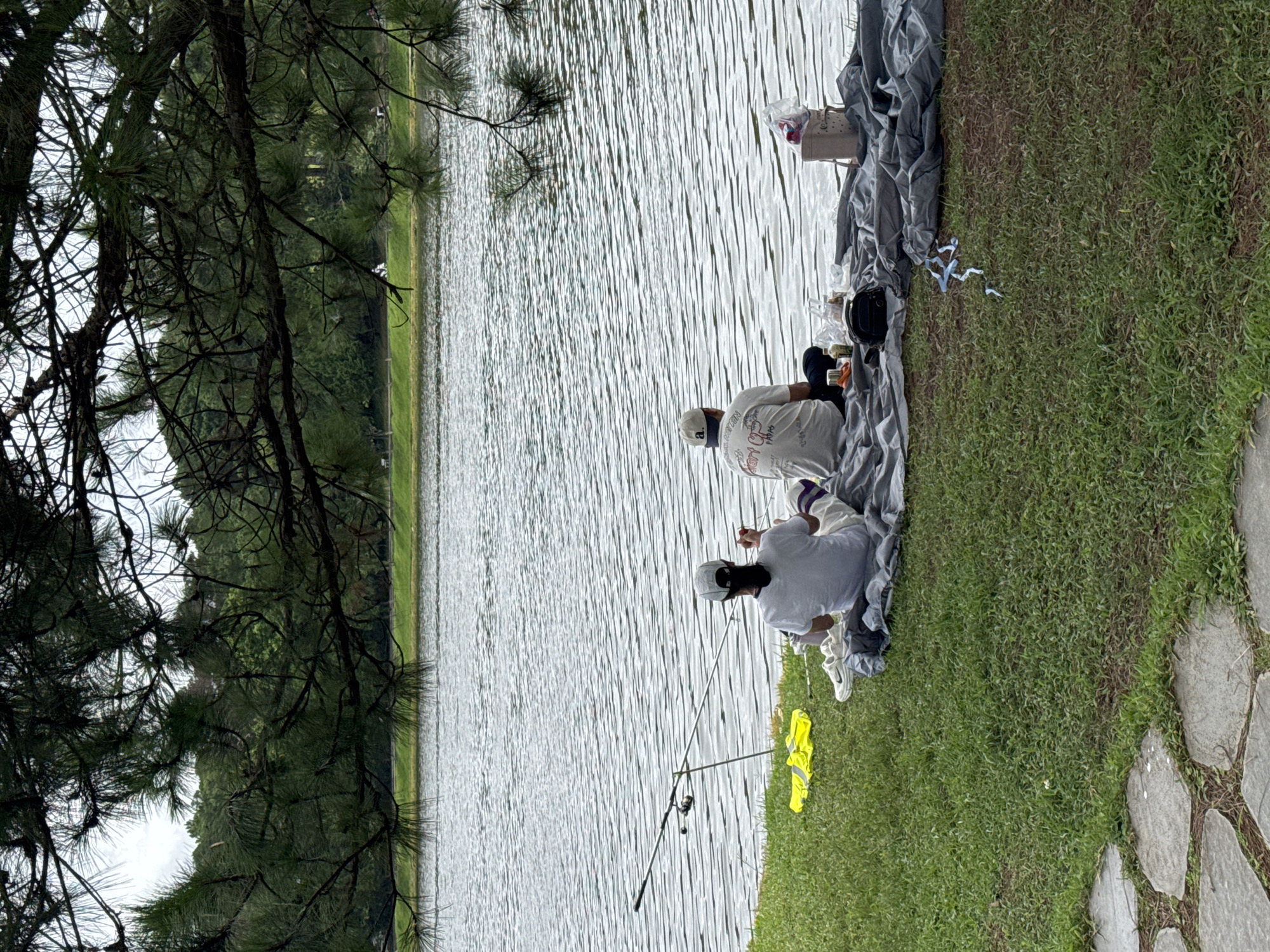 Two individuals are picnicking by Xuan Huong Lake in Da Lat, Vietnam. The woman on the left, wearing a white t-shirt and a white cap, is seated on a gray blanket with a yellow vest to her left. The man on the right, wearing a white t-shirt with graphics and a white cap, is also seated on the blanket. A picnic basket, food containers, and a camera bag are spread on the blanket. A fishing rod is set up near the woman, suggesting they may also be fishing. The scene is set against the backdrop of the calm, rippling waters of Xuan Huong Lake, with lush green grass and trees forming the foreground and middle ground. The image is framed by the dark branches of pine trees in the upper portion, casting a shadow over the scene.