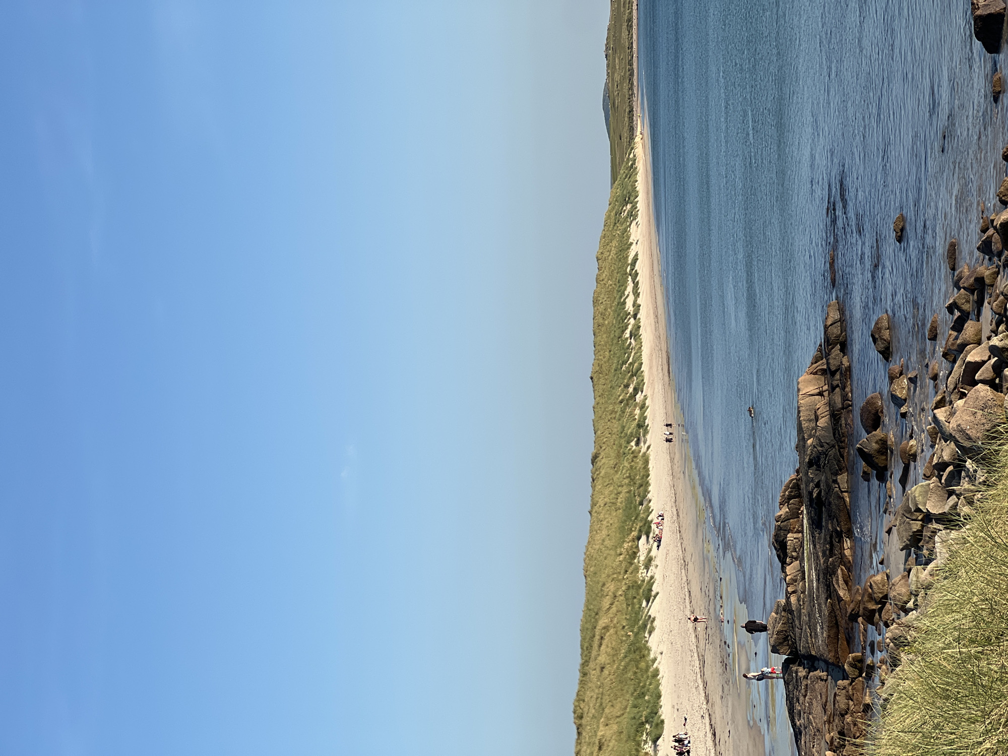 This is a wide-angle, eye-level shot of a sandy beach with dunes and a calm ocean under a clear blue sky in Lunniaghbeg, Ireland. The foreground is dominated by rough, dark-colored rocks covered in patches of green grass. In the mid-ground, the beach is populated by several individuals and small groups of people, some appearing to be sunbathing or relaxing, while others are walking along the shore or in the shallow water. A dog is visible in the water. The sand gives way to a gentle slope of grassy dunes that stretch across the background. The ocean is a deep blue with slight ripples. In the far distance, a low, green hill is visible on the horizon. The lighting suggests it is late afternoon or early evening.