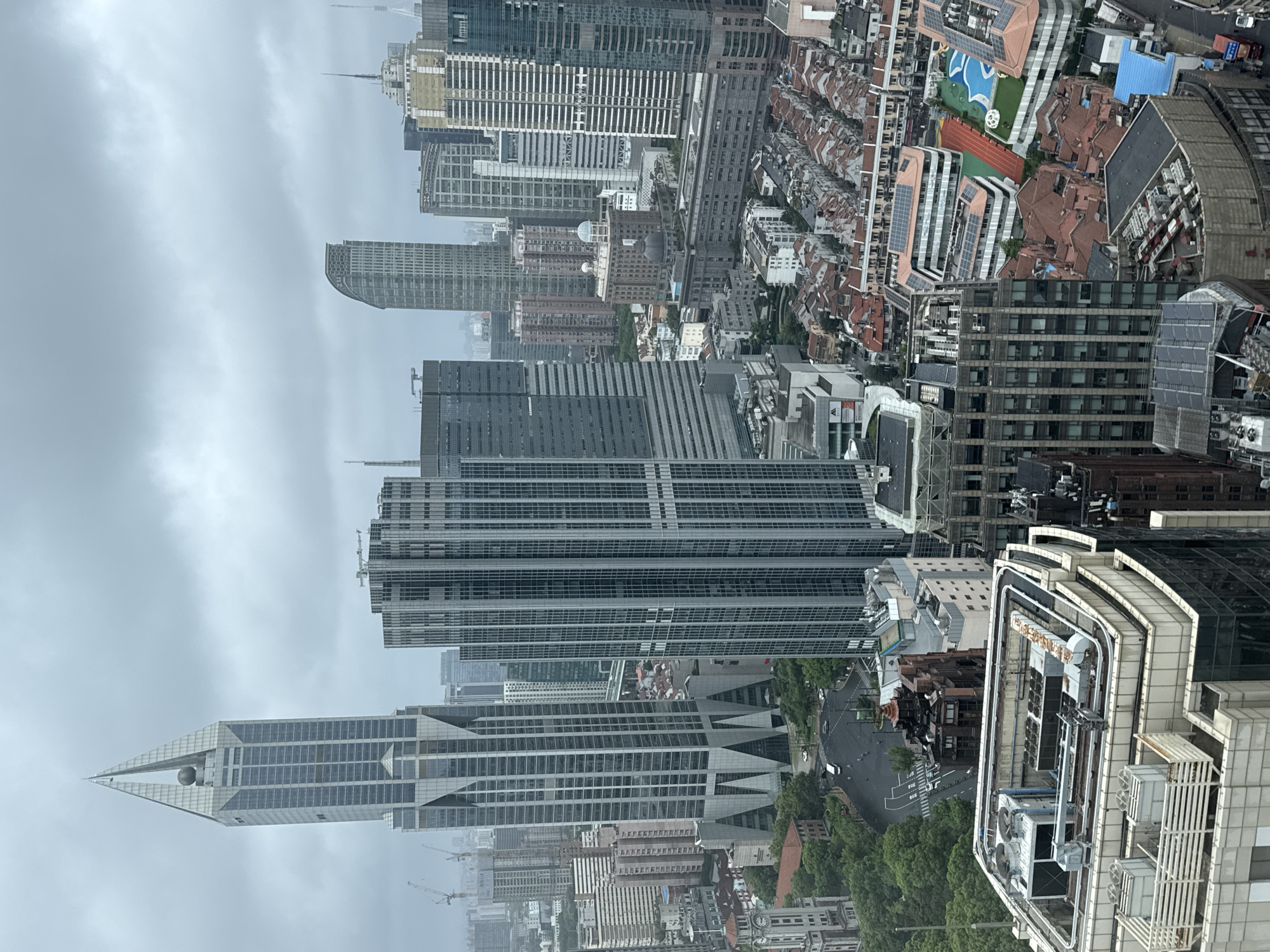 This is a high-angle, eye-level, medium shot of a dense urban skyline in Shanghai, China, under an overcast sky. The primary focus is on a collection of modern skyscrapers of varying heights and architectural styles. Dominating the left side of the image is a very tall, slender skyscraper with a pointed, faceted apex that resembles a diamond or a shard of glass. Its facade is primarily glass and metal. To its right, a wider, rectangular skyscraper with a grid-like facade is prominent. Further into the background, a variety of other tall buildings, some with curved facades and others with more traditional rectangular forms, can be seen. Interspersed among the modern structures are lower-rise buildings with red tile roofs, characteristic of older residential areas, some of which appear to have been redeveloped with modern additions, including solar panels. A swimming pool and a running track are visible within one of these complexes. The street level is not clearly visible, but glimpses of roads, vehicles, and trees suggest a busy urban environment. Construction cranes are visible in the far distance, indicating ongoing development. The overall impression is one of a rapidly modernizing metropolis with a mix of historical and contemporary architecture.