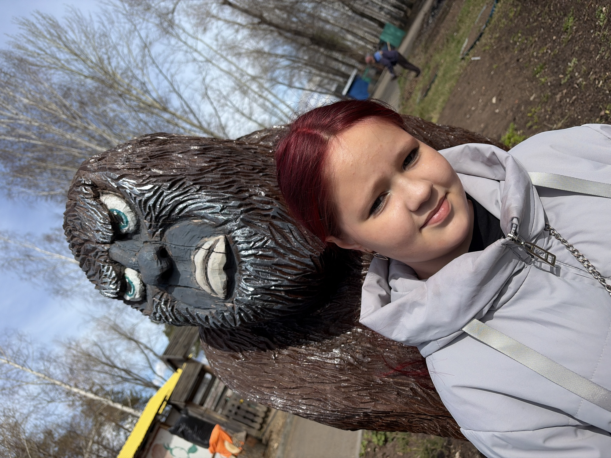 The image presents a vibrant outdoor scene featuring a young person and a large, carved wooden statue, set against a backdrop of bare trees and a clear sky. In the foreground, a young person with striking red hair is shown smiling warmly towards the camera. She is wearing a light grey puffer jacket, and a chain strap is visible on her right shoulder.
To the left of the young person is a detailed wooden statue, resembling a gorilla or Bigfoot, with carved brown fur, distinct green eyes, and bared white teeth, giving it a playful yet imposing presence.
The background reveals a natural environment, possibly a park or recreational area. Numerous deciduous trees with bare branches indicate a season of late winter or early spring. The ground is a mix of dry earth and sparse vegetation. In the mid-distance, another individual is seen bent over, engaged in an activity on the ground, wearing a blue top and dark trousers. The bright daylight suggests it is either morning or afternoon. The overall atmosphere is pleasant and casual, with the young person appearing happy and relaxed. No visible text is present in the image. The setting aligns with an outdoor location in Chaikovsky, Russia.