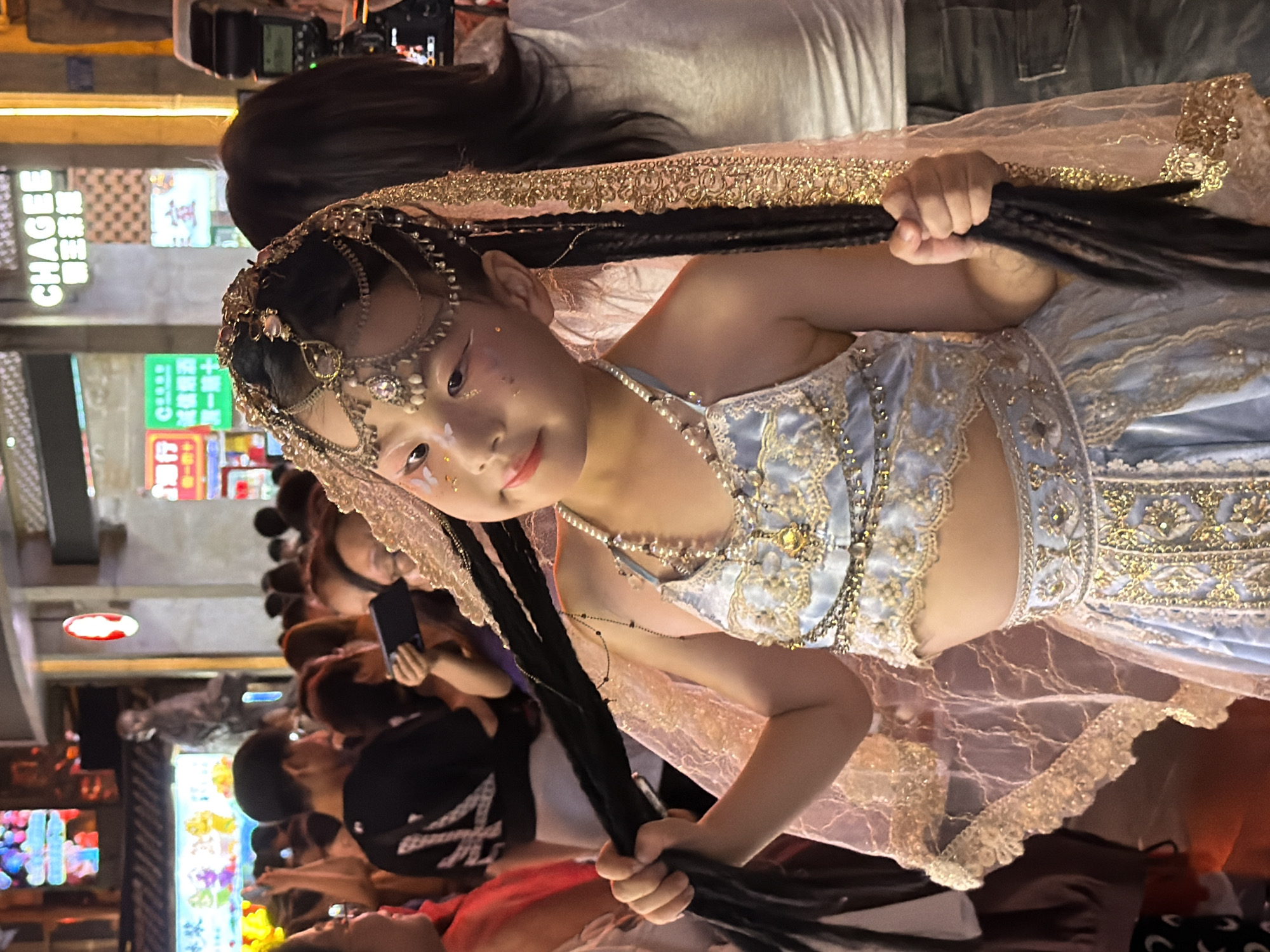 The image captures a young girl dressed in an elaborate, light blue and gold costume, possibly a traditional outfit or for a performance. She is adorned with intricate jewelry, including a jeweled headdress, earrings, and necklaces. Her hair is styled in long braids, and she is holding a portion of her hair in her hand. The costume features ornate embroidery and delicate lace details.
The scene appears to be an outdoor setting at night, given the artificial lighting from storefronts and signs. The background suggests a bustling street or market area, with blurred figures of other people and various illuminated displays. Some of the visible text in the background includes "CHAGEE" on a sign, and other characters that are difficult to decipher due to the blur and lighting.
The young girl's expression is somewhat neutral, with a hint of a smile. The overall atmosphere seems to be one of a public gathering or event, possibly a festival or a street performance, given the elaborate costume and the presence of onlookers. The lighting creates a dramatic effect, highlighting the girl and her attire against the darker background. The image has a slightly tilted perspective.