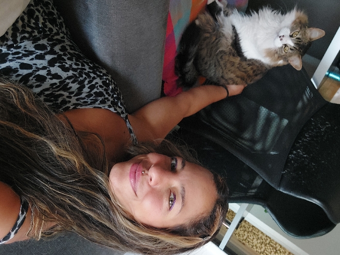 A woman with long, highlighted brown hair and a lip piercing reclines on a gray couch in a sleeveless black and gray patterned top. She holds out her arm, upon which rests a fluffy, long-haired cat with brown, black, and white markings. The cat is reclined on a black mesh office chair. The background features a brightly lit room with hints of white furniture and a colorful throw pillow on the couch.