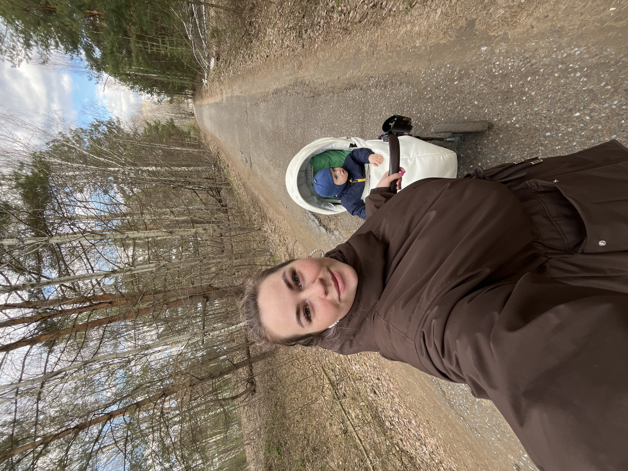 A woman captures a selfie while walking along a forest path with a child in a stroller. The image is taken from a first-person perspective, with the woman's face and upper body visible in the foreground, angled to the right. She has light-colored hair, dark eyebrows, and wears a brown puffer jacket, exhibiting a slight, pleasant smile. Her left hand, with light pink nail polish, holds what appears to be the stroller handle or the camera.
Behind and to the left of the woman, a young child is seated in a white, modern stroller. The child wears a blue hat and a dark jacket over a green layer, looking calmly towards the camera.
The setting is a narrow, unpaved path made of dirt and gravel, winding through a wooded area. The path is flanked by numerous trees, including bare deciduous trees with white bark (likely birches) and some evergreen conifers (pines or firs). The bare branches suggest late autumn, winter, or early spring. Above the trees, patches of blue sky with white clouds are visible. The scene depicts a serene outdoor stroll, likely in Chaikovsky, Russia, during cool but fair weather conditions. No text is visible in the image.