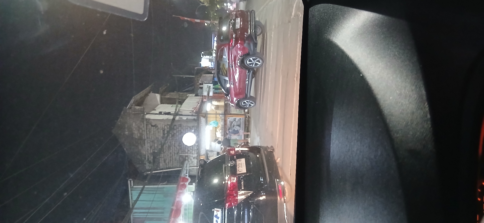 At night, on a road in Samarinda, Indonesia, a black SUV is parked. The SUV is facing right, and its license plate reads "KT 1434 AN." To the right of the black SUV, a red SUV is also parked and facing right. This red SUV has a license plate that is partially obscured. In the background, there is a building with a brick facade and multiple levels. There are also several stalls or shops with awnings. The sky above is dark, with visible power lines. The road surface has white stripes, suggesting a crosswalk or lane markings. The foreground is dominated by the dark interior of what appears to be a vehicle, possibly a dashboard and part of a windshield or interior mirror.
