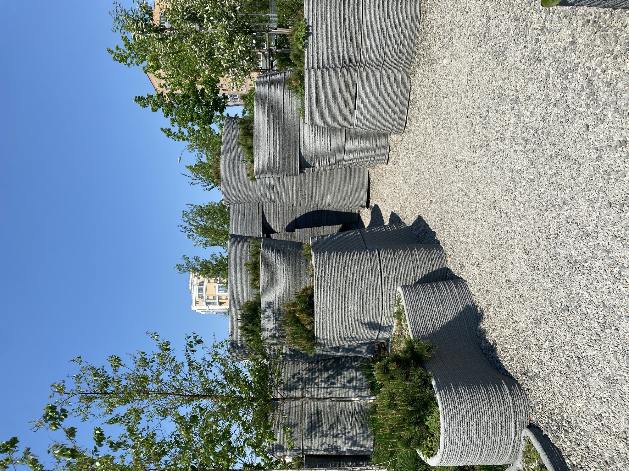 The image depicts a unique urban landscape in Yekaterinburg, Russia. The scene consists of a series of vertically stacked, gray, rope-like structures, resembling planters or seating areas. The structures appear to be constructed using a layered technique, giving them a textured, ribbed appearance. Each tier of the structures is filled with various green plants and trees. The ground is covered in a light-colored gravel or pebble material. The background features a clear blue sky, green trees, and a multi-story building in the distance. The lighting suggests a sunny day.