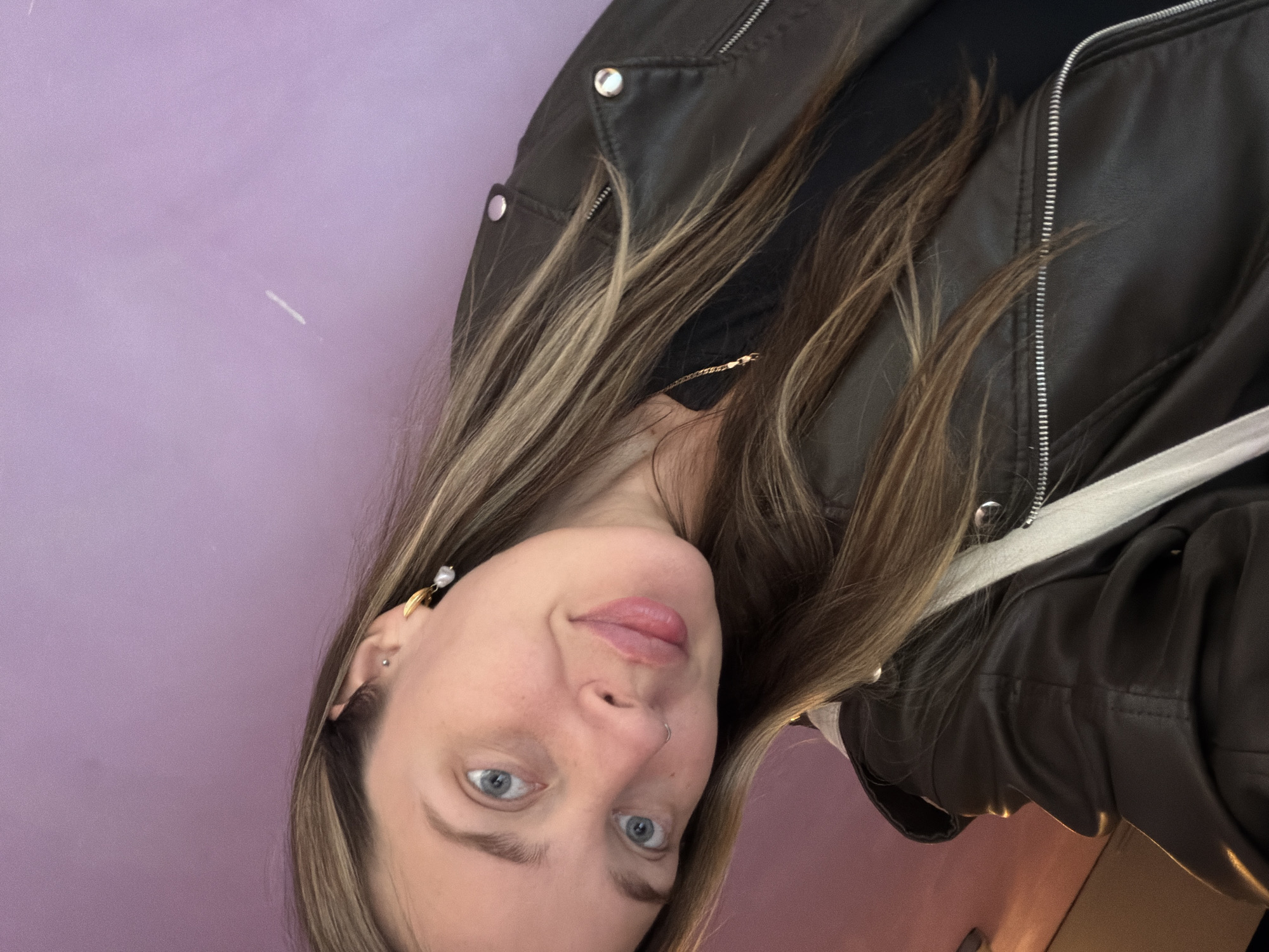 A woman with blonde and brown hair, blue eyes, and a nose piercing is the primary subject of this close-up, slightly angled photograph. She wears a dark leather jacket over a black top, and her jewelry includes earrings with pearl accents and a thin gold necklace. The background is a pale purple wall, seemingly indoors. The location in the city of Chaikovsky, Russia, is not discernible from the image.