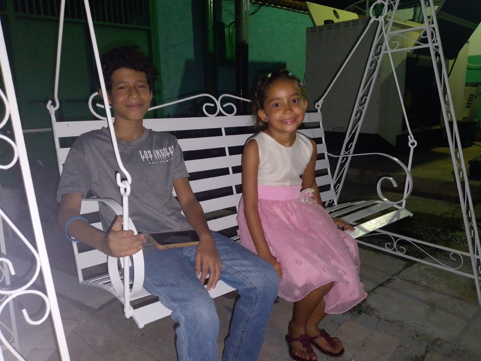 Two children, a boy and a girl, are sitting on a white metal swing set. The boy, wearing a grey t-shirt, jeans, and a blue bracelet, is holding a mobile phone. The girl is wearing a white top and a pink dress with brown sandals. The location appears to be outside in Tinaquillo, Venezuela. A building and a light fixture are visible in the background.