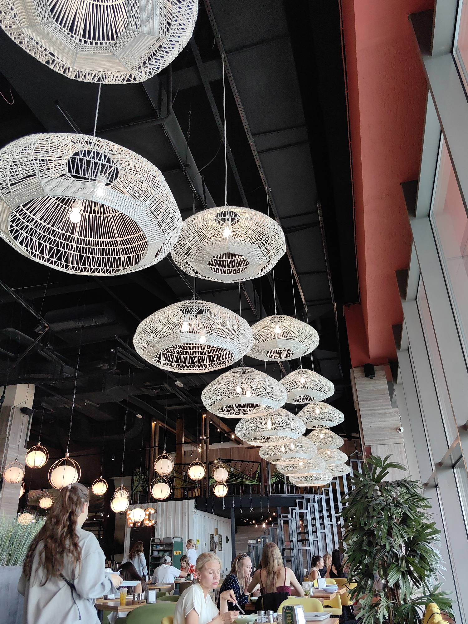The image shows the interior of a modern, dimly lit restaurant with a focus on overhead lighting fixtures. A series of large, white, woven pendant lamps hang from the dark ceiling, casting a warm glow. Several smaller, globe-shaped pendant lights are also visible in the background. The walls are a mix of dark surfaces and a vibrant red accent wall. Large windows line the right side of the frame, revealing an outdoor scene or another part of the building. Inside, tables are set with chairs, and several patrons are seated and dining. A woman with long, dark, wavy hair is in the foreground, facing away from the camera, seated at a table. Another woman with blonde hair is visible at a table to the right, looking down. A lush green plant is situated in the lower right corner of the image. The restaurant appears to be busy, with staff and patrons mingling. The overall atmosphere is contemporary and inviting.