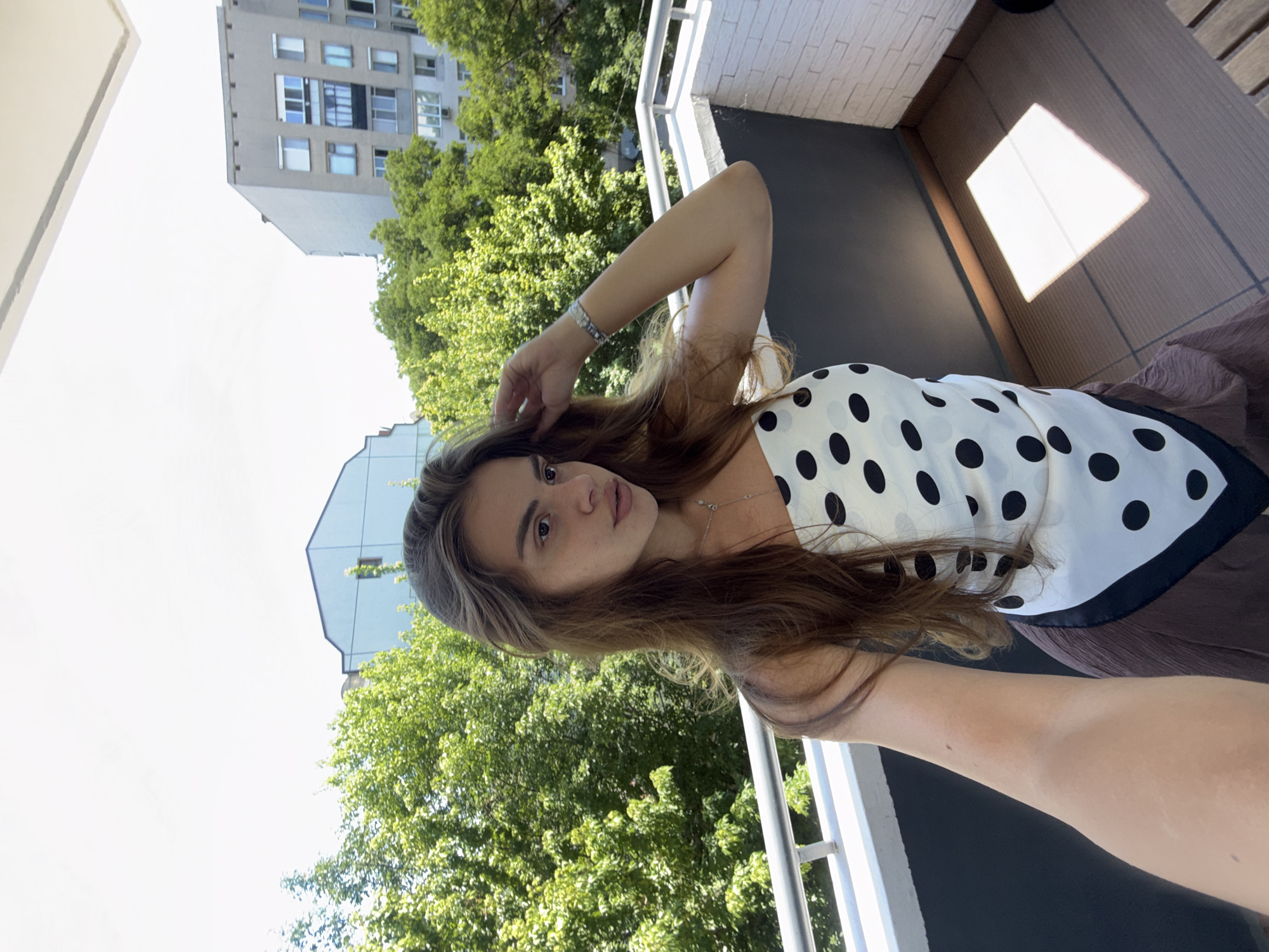 A young woman with long brown hair takes a selfie on a balcony in the city of Plovdiv, Bulgaria. She is wearing a white top with black polka dots and a brown skirt. The balcony overlooks a street lined with trees and apartment buildings. The sky is clear and blue. The woman is smiling and looking at the camera. She is wearing a silver bracelet on her left wrist and a delicate silver necklace. Her right hand is touching her hair. The balcony floor is made of wood.