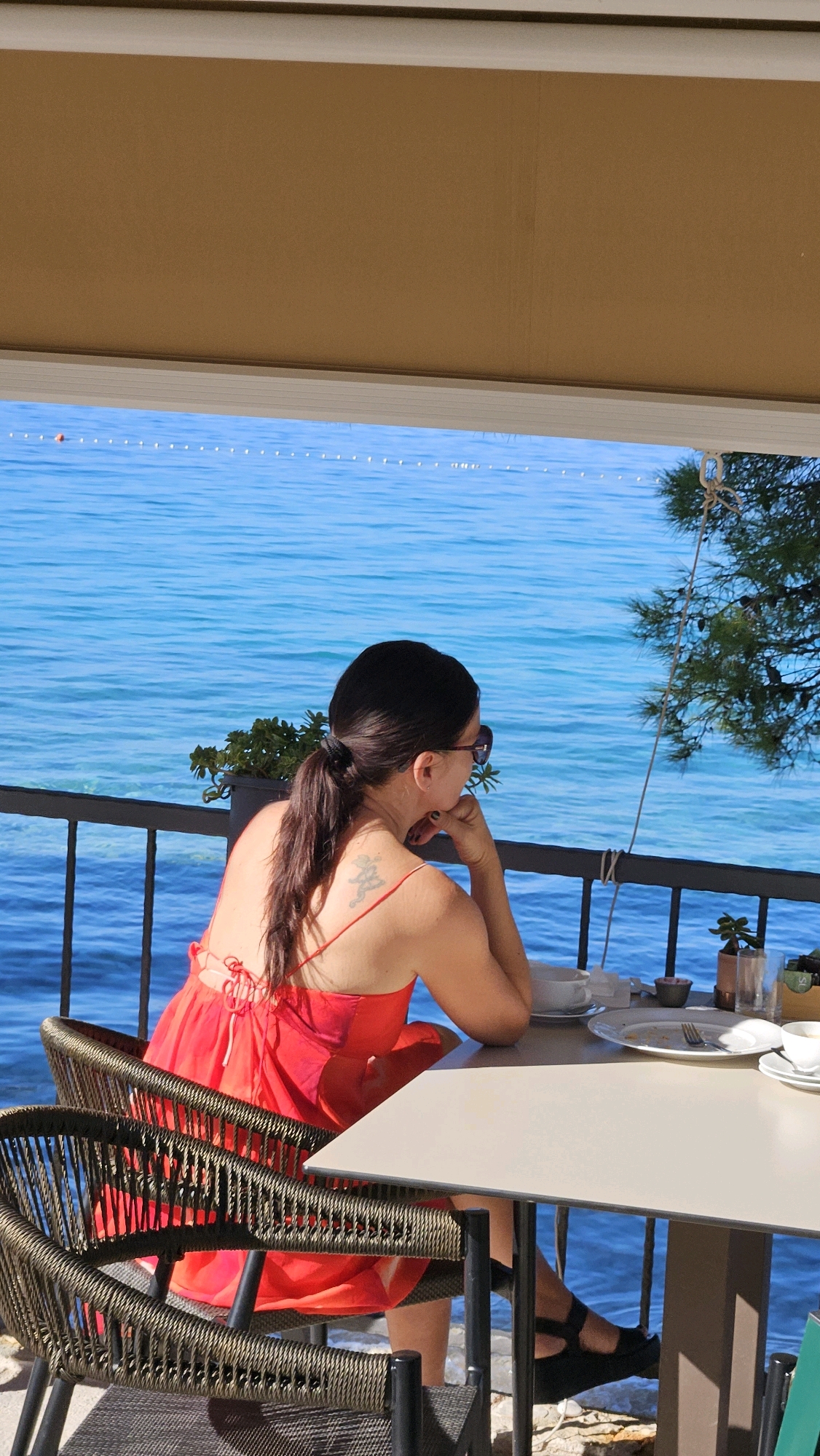 A woman with dark hair in a ponytail, wearing sunglasses and a red dress, sits at a table on a balcony overlooking the sea in Općina Tučepi, Croatia. She is resting her chin on her hand, looking out at the water. On the table are plates, cups, and cutlery, suggesting she has recently finished a meal. The balcony has a railing, and a potted plant sits near the woman. The sea is a vibrant blue, and a line of buoys is visible in the distance. Sunlight filters through an awning above, casting shadows on the scene.
