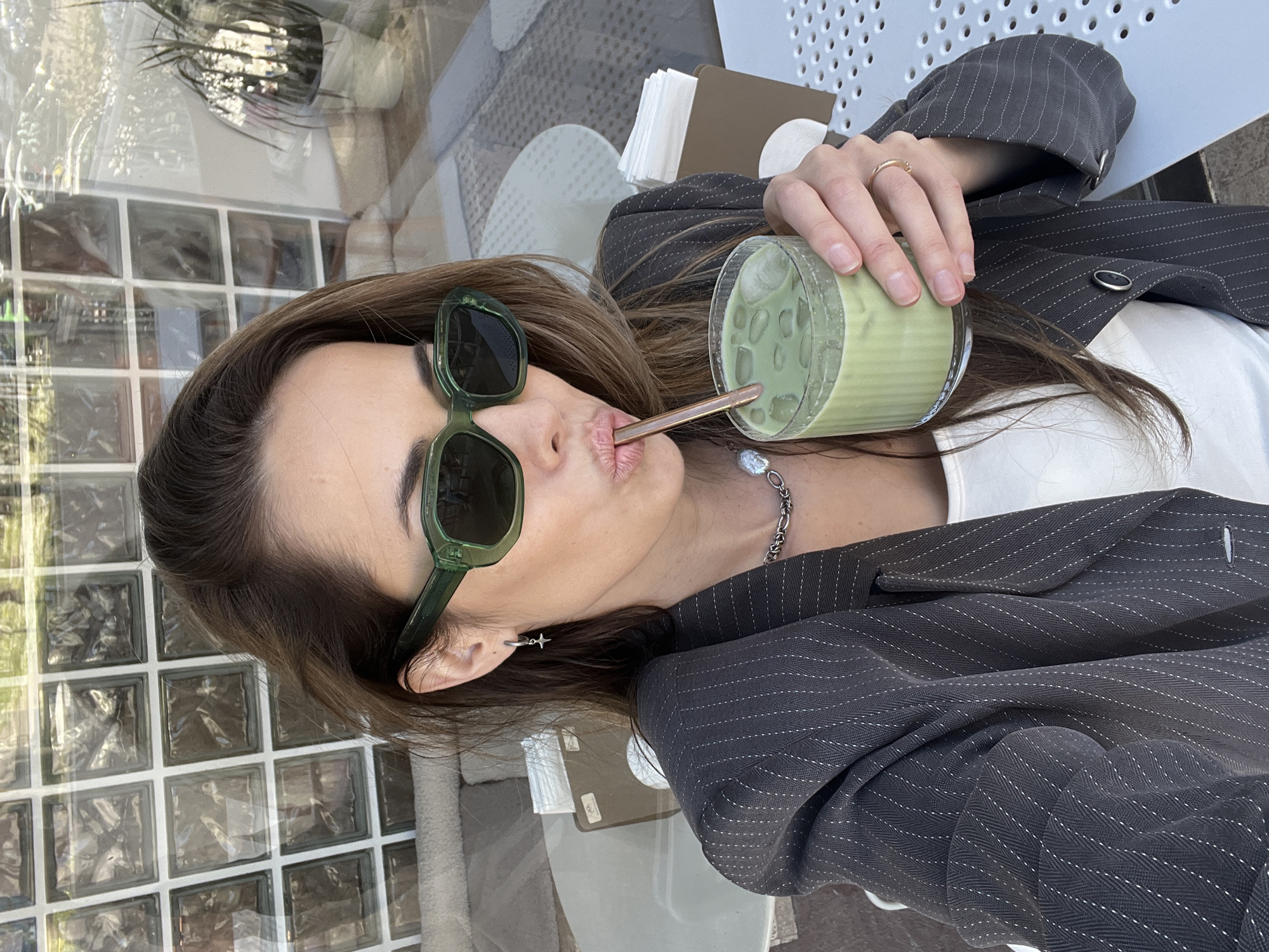 A medium-close-up photograph shows a woman seated at an outdoor cafe drinking a beverage, possibly a matcha latte, through a straw. She wears dark green sunglasses and a gray pinstripe blazer over a white top. She has a silver chain necklace and a star earring in her left ear. Her brown hair is parted on the side. The cafe appears to be in Bar, Montenegro. The table is a light color, and items such as napkins and a card or small notebook are present. The background includes a glass block wall.