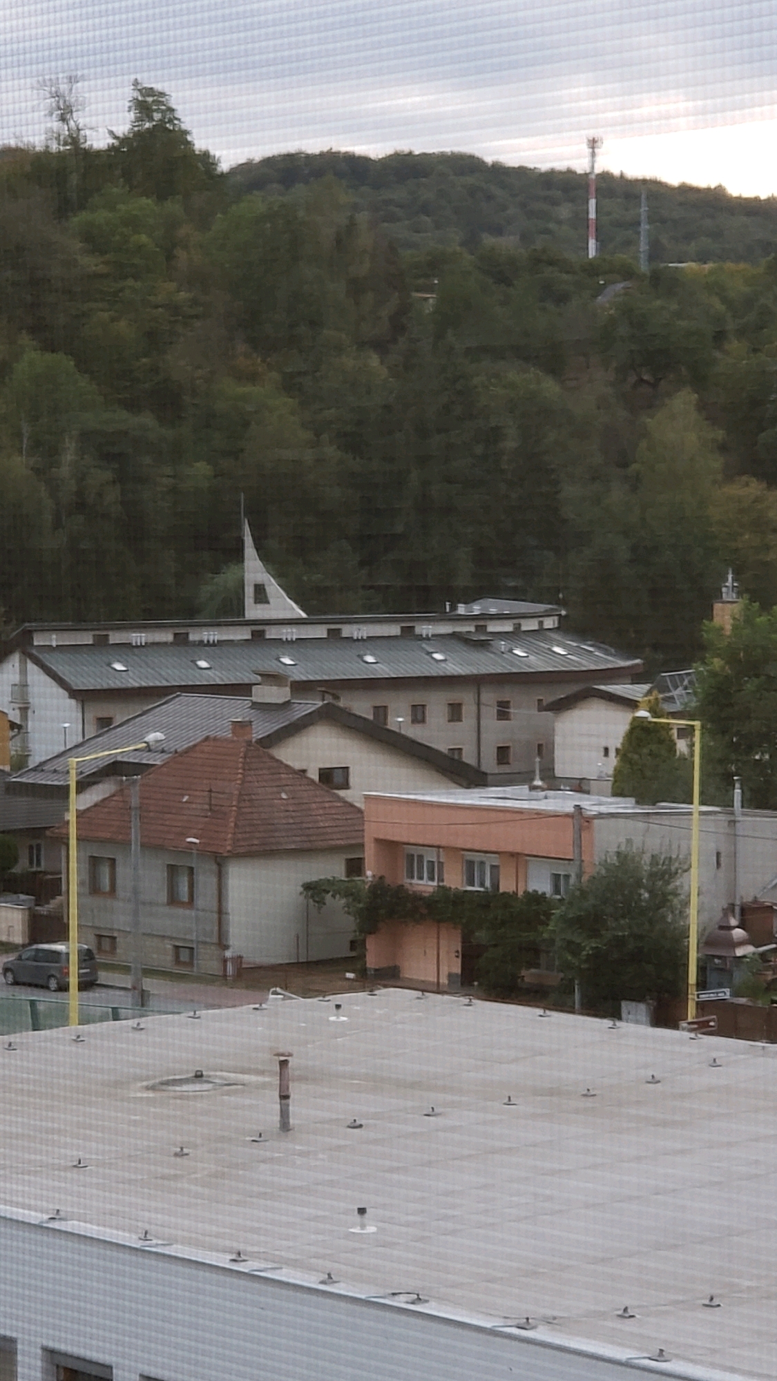 The image shows a residential area with several buildings of varying heights. In the foreground, a flat-roofed building with a white facade and visible fasteners on the roof is prominent. A few ventilation pipes and small antenna-like structures dot this roof. Behind it, a two-story building with a red-tiled roof and beige walls is visible, featuring several windows. To its right, a light orange building with a flat roof and multiple windows stands. Further back, a larger, elongated building with a dark roof and numerous skylights is present, and a distinct white, triangular spire rises above it. The background is dominated by a dense forest covering rolling hills. A telecommunications tower with red and white bands is visible on the crest of the hill in the upper right portion of the image. A dark-colored car is parked on the street in front of the red-roofed building. The overall scene suggests a suburban or semi-rural environment.