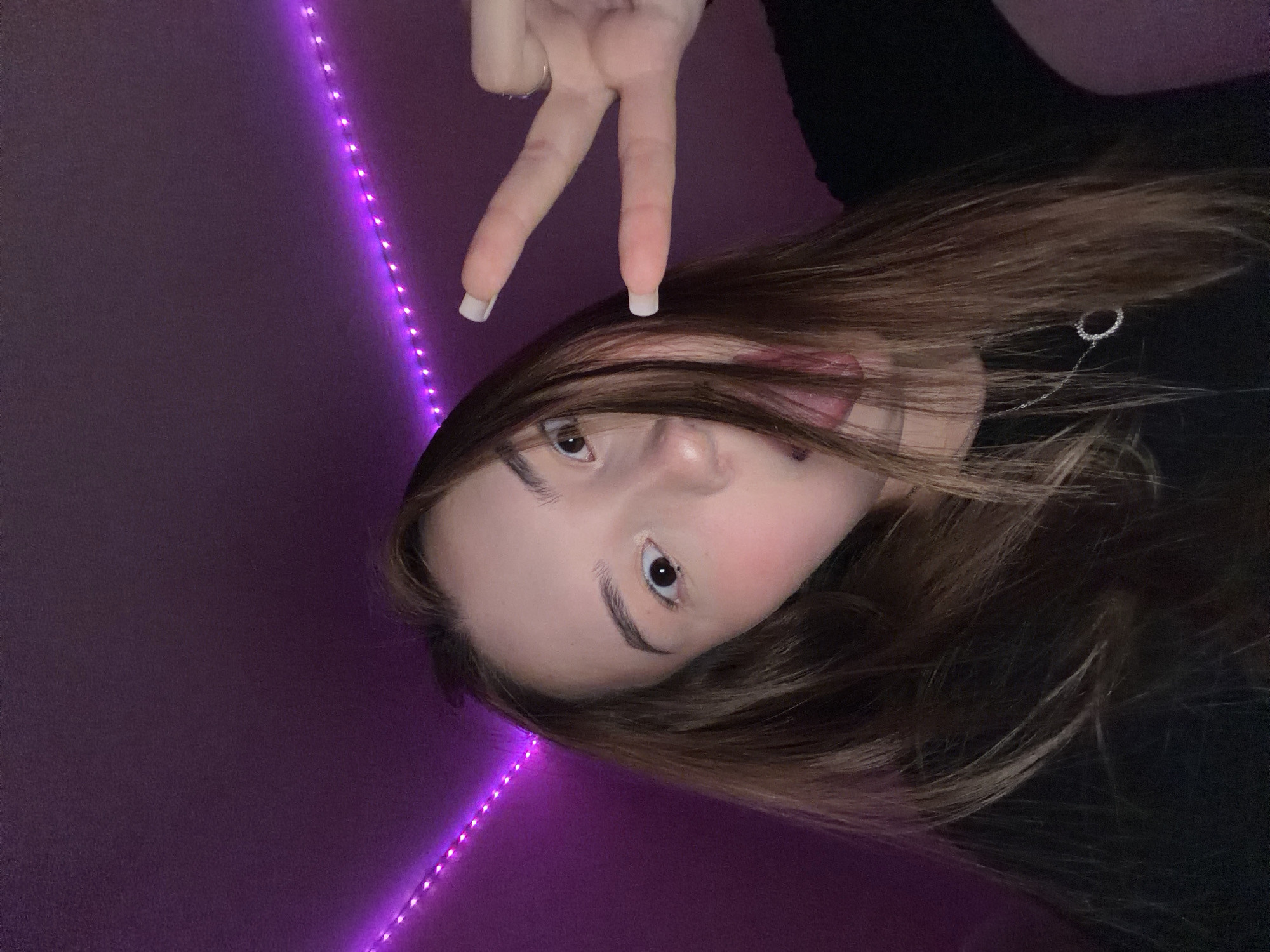 A young woman with brown hair is making a peace sign with her fingers. She is indoors against a purple wall with a string of purple LED lights. The location is unknown and there is no evidence to suggest she is in Chaikovsky, Russia.