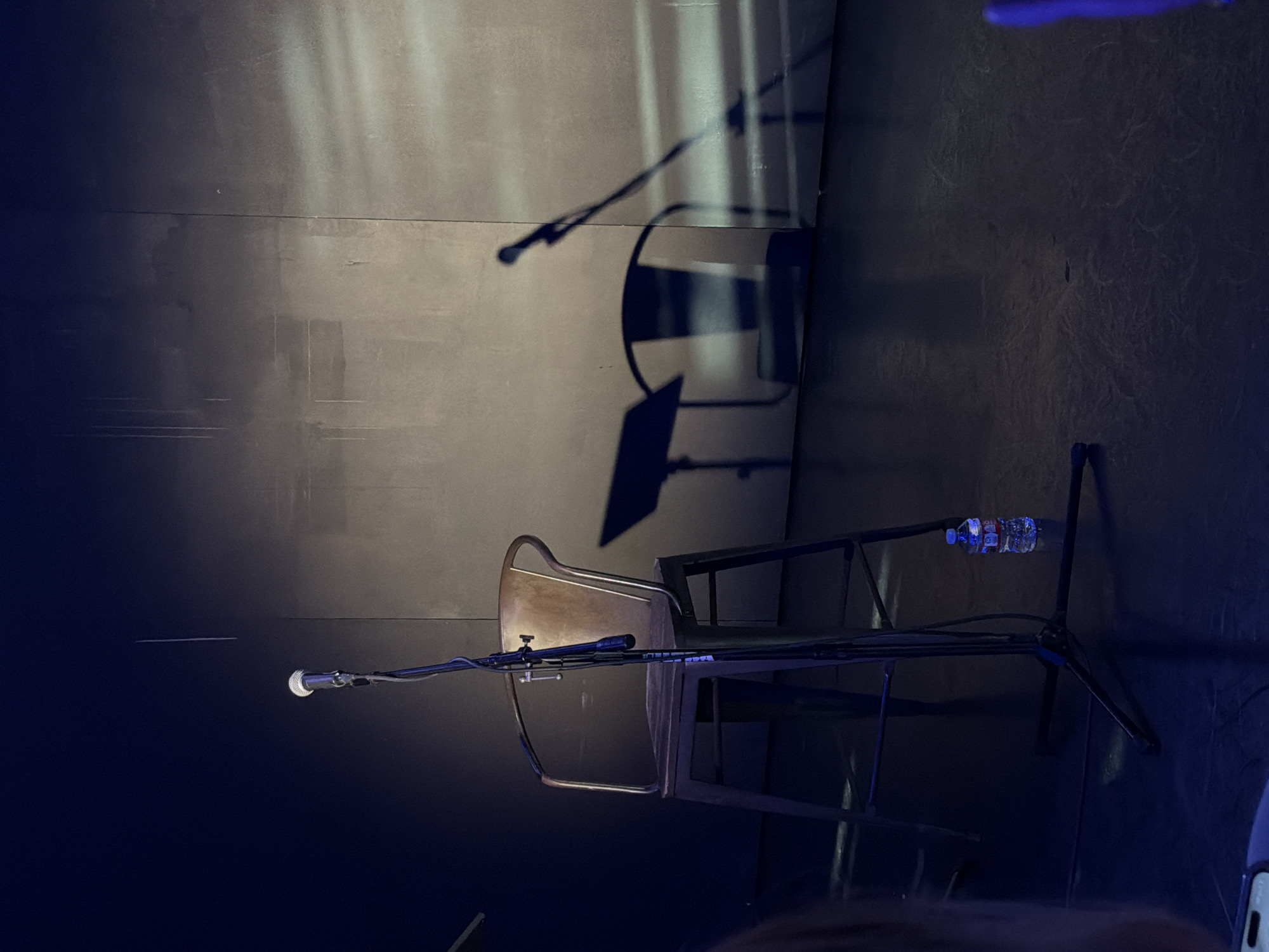 The image depicts a stage setup with a single stool, a microphone on a stand, and a water bottle on the floor. The stool is made of metal with a weathered finish and has a minimalist design with a backrest and armrests. The microphone is positioned in front of the stool, and its shadow is cast onto the textured wall behind it, along with the shadow of the stool and a music stand. The wall appears to be a dark, possibly concrete surface, with subtle lighting effects creating a dramatic atmosphere. The floor is dark and reflective, indicating it might be a stage floor. The overall lighting is dim, with a spotlight creating a strong contrast between illuminated areas and shadows. No people are visible in the frame, suggesting the scene is set before or after a performance. The composition focuses on the emptiness of the stage, emphasizing the anticipation or aftermath of an event. The specific location of this scene within Los Angeles, United States of America, cannot be determined from the provided image alone.