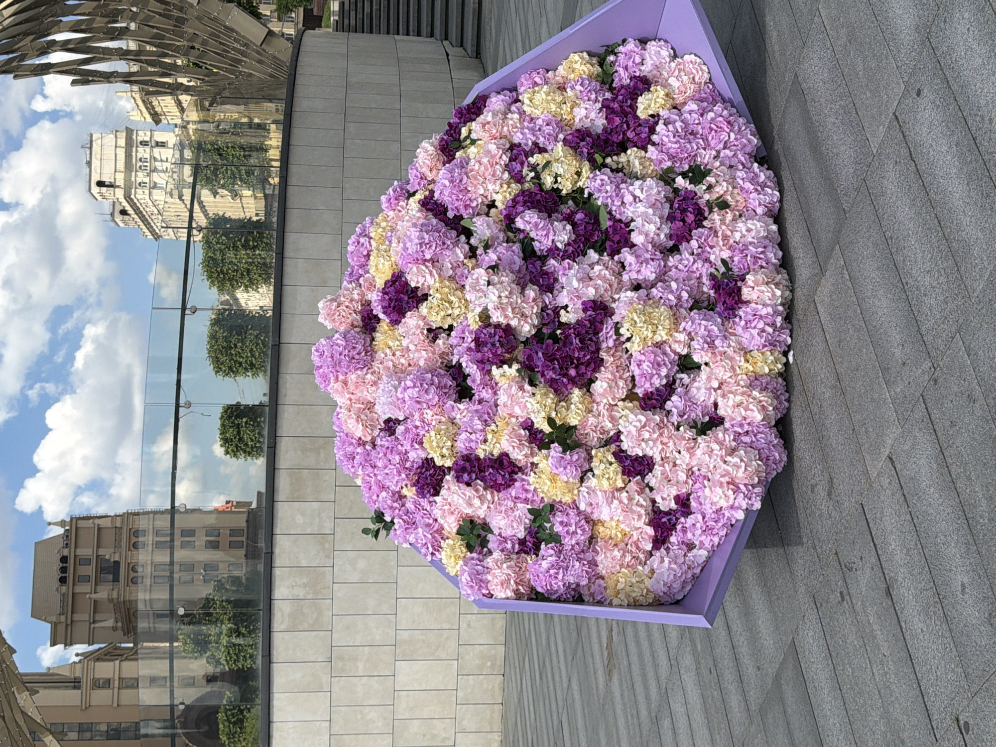 The image depicts a vibrant heart-shaped floral arrangement placed outdoors in a public space in Moscow, Russia. The arrangement is composed of numerous artificial flowers in shades of light purple, dark purple, pink, and pale yellow, creating a dense, textured surface reminiscent of hydrangeas. It is housed in a low, open-top container of a solid lavender or light purple color.
The floral display rests on a paved ground with grey, rectangular tiles. To the left (after rotating the image to the correct orientation), there is a section of a beige-tiled pathway. In the background, a modern architectural element with angled metallic slats is visible. A transparent glass barrier runs horizontally across the mid-ground, reflecting a bright blue sky with white clouds, as well as parts of a grand, classical-style building with multiple windows and decorative elements. Further reflections show green foliage from trees and other urban structures. Beyond the glass, parts of other buildings and trees are visible, confirming an urban setting.
The scene is brightly lit by natural sunlight, suggesting it is daytime with clear skies. No people are visible, and no specific activity is taking place other than the static display of the floral arrangement. There is no visible text in the image. The overall impression is one of an artful and celebratory urban decoration.