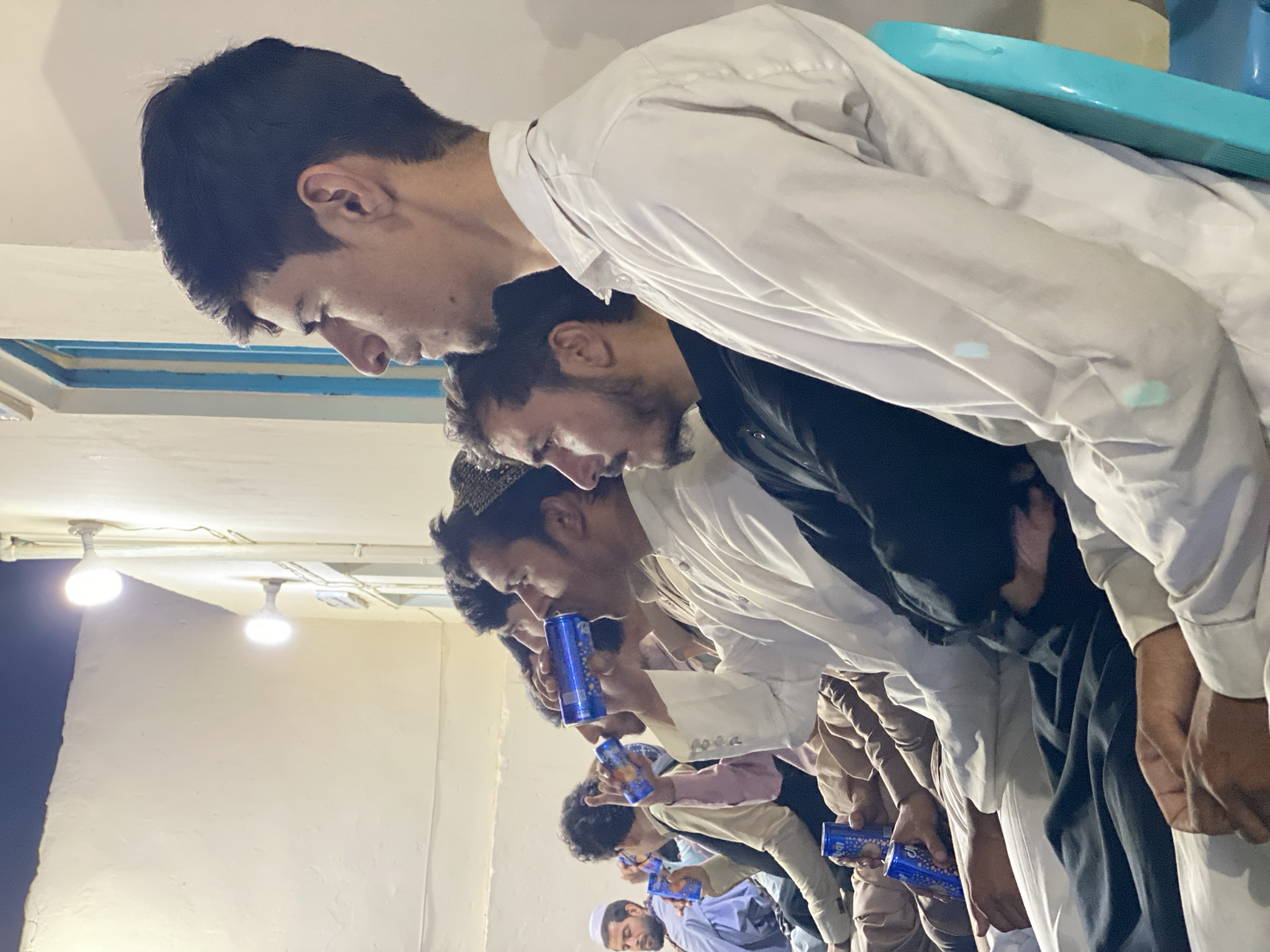 The image depicts a group of men sitting in a row, facing right, in what appears to be an outdoor setting at night, likely in Kandahar, Afghanistan. The lighting suggests artificial illumination from wall-mounted lights. Several men are holding and drinking from blue cans, some of which have a patterned design. The men are dressed in traditional clothing, including shalwar kameez and caps. One man in the foreground, wearing a white shalwar kameez, is holding a blue can to his mouth. Another man to his left, also in a white garment, is drinking from a similar can. Further to the left, another man is seen holding a blue can. The background consists of a textured wall and a blue-trimmed doorway or window. The overall atmosphere is informal, with the men appearing relaxed as they engage in conversation or observe something out of frame.