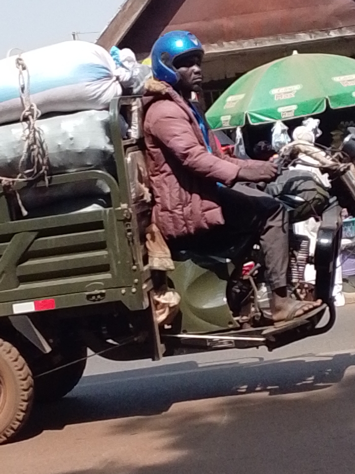 An adult male with dark skin is operating an olive-green three-wheeled motor-tricycle, commonly known as an "Aboboyaa," on a paved road in a commercial or market district within the city of Unknown, Ghana. He is wearing a blue full-face helmet, a reddish-brown padded jacket over a blue garment, dark trousers, and open-toed sandals. His hands are positioned on the handlebars, and his gaze is directed forward. The vehicle's cargo bed, positioned to the left of the driver, is loaded with multiple large, white or light-colored sacks, secured by ropes. One prominent sack has a bluish tint. A thick coil of rope hangs from the left side of the cargo bed. In the background, to the right, a large green market umbrella with visible white text is present, alongside indistinct building structures. The scene is illuminated by strong daylight, casting prominent shadows beneath the vehicle.