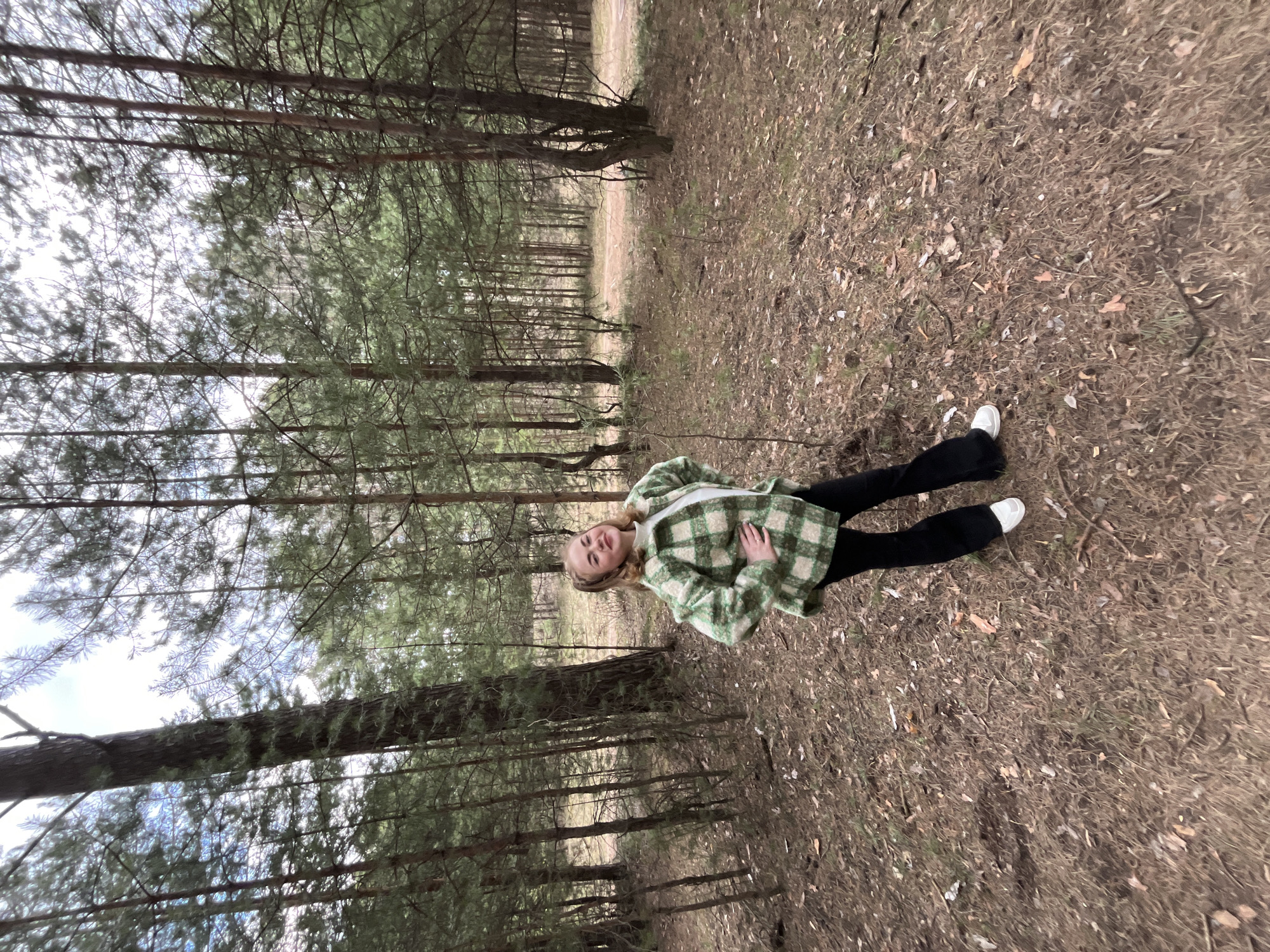 A young woman is depicted lying on her back on the forest floor, looking up at the camera with a gentle smile. She has blonde hair and is wearing a green and white plaid shacket or coat over a white top, black pants, and white sneakers. Her left hand rests on her torso.
The setting is a dense pine forest. Tall, slender pine trees with sparse branches line the background, with the sky visible through the canopy, suggesting an overcast day. The ground is covered with dry pine needles, fallen leaves, and sparse brown grass, indicating a period like late autumn, winter, or early spring. A narrow, unpaved path or clearing can be seen further back among the trees. The overall mood is calm and natural.
The activity taking place is a posed photograph in an outdoor setting. The time of day is daytime, with soft, diffused lighting. The location is a forest in Chaikovsky, Russia. There is no visible text in the image.