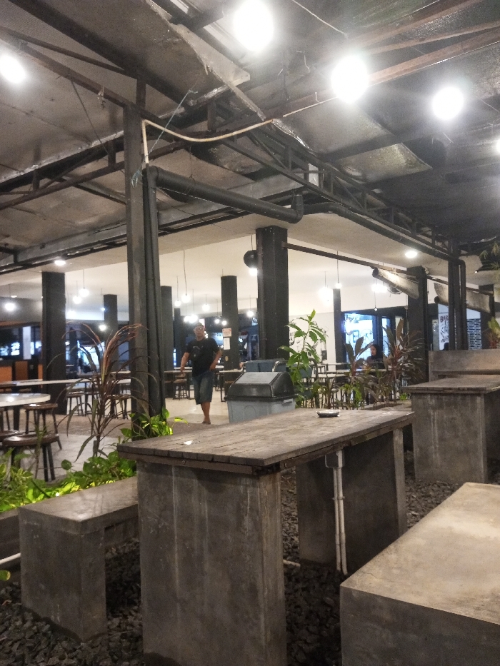 The image displays an indoor or semi-open-air commercial dining space within an establishment in Pontianak, Indonesia. The ceiling features an exposed industrial aesthetic with black metal trusses, large black pipes, and multiple bright, circular overhead light fixtures. The floor is covered with dark grey gravel. Seating consists of numerous tables and accompanying benches constructed from unpolished concrete, each topped with a dark wooden slab.
In the mid-ground, a male individual, wearing a dark short-sleeved shirt and dark shorts, walks barefoot from the right towards the left side of the frame. To his right, a light grey lidded trash bin is visible. Various green and red-hued potted plants are interspersed throughout the seating area, defining sections. On the wooden surface of the large concrete table in the foreground, a small white ashtray is present. White electrical conduits are attached to the concrete supports of some tables. Multiple dark-colored structural pillars are spaced across the area. In the background, additional tables and chairs are visible, with several indistinct figures seated, suggesting active patronage. The scene is evenly illuminated by the overhead lighting.
