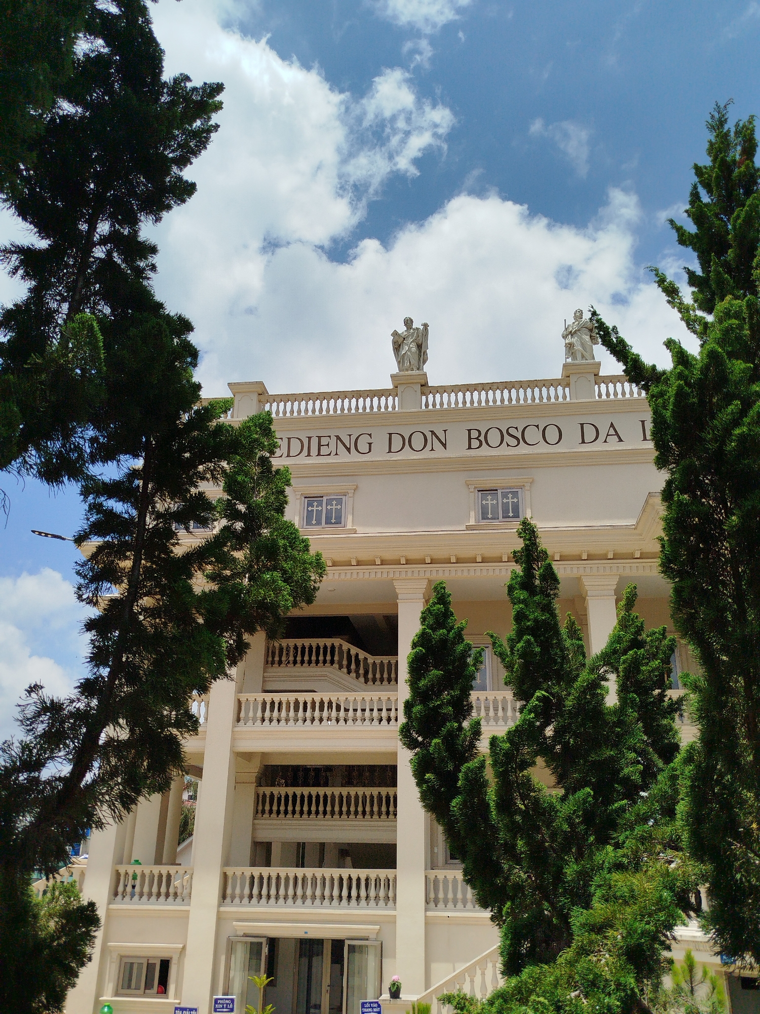 The image displays a grand, light-colored building, likely a religious or educational institution, partially obscured and framed by several tall, dark green coniferous trees. The building features classical architectural elements, including prominent white columns supporting upper floors and ornate white balustrades on its balconies and roofline.
On the roof, two white statues of religious figures are visible, one appearing to hold a book or scroll, and the other a staff or scepter. A prominent sign spanning the building's facade clearly reads "EDIENG DON BOSCO DA L", indicating a Salesian institution, possibly "Thánh Điện Don Bosco Đà Lạt" (Shrine of Don Bosco Da Lat) or a similar center in Da Lat, Vietnam, as per the provided context. One window on the main facade notably features a white cross motif.
The scene is captured on a bright day, under a blue sky adorned with scattered white, fluffy clouds. The sunlight illuminates the building and trees, suggesting a time around midday or early afternoon. No discernible human activity or event is taking place, presenting a static yet impressive architectural view surrounded by nature. A black streetlight is partially visible through the trees on the left side of the frame.