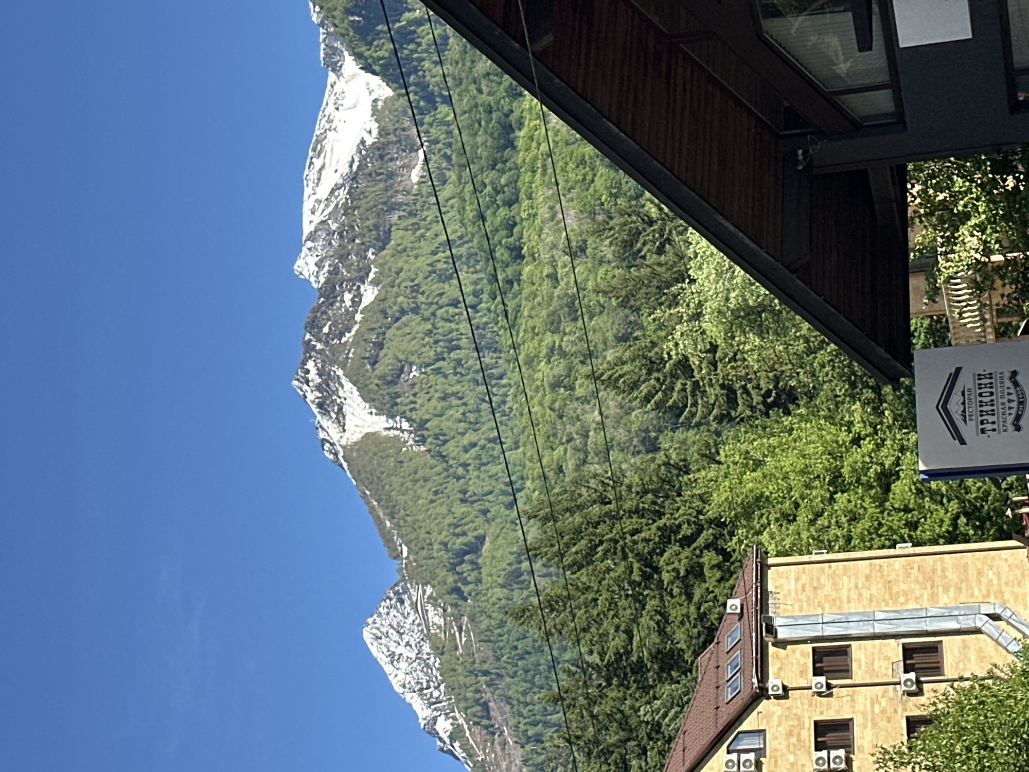 This image captures a panoramic view of a majestic mountain landscape interspersed with modern architecture, likely within a resort setting. The scene is dominated by towering mountains, whose upper slopes are still covered in patches of snow, contrasting sharply with the deep green of dense coniferous forests that blanket the lower and middle sections of the peaks. A clear, vibrant blue sky occupies the upper left portion, indicating bright, sunny weather, possibly midday.
In the foreground, on the right side of the image, man-made structures are visible. Part of a contemporary building with a dark facade and a distinctive wooden underside or soffit extends into the frame from the top right. Attached to this structure is a prominent white sign with black Cyrillic text. The text reads "РЕСТОРАН" (RESTAURANT) at the top, followed by "ТРИКОНИ" (TRIKONI) as the main name, and "КРАСНАЯ ПОЛЯНА" (KRASNAYA POLYANA) at the bottom, identifying a restaurant in the specified location.
Below this sign and further to the right, a multi-story building with a light-colored, possibly beige or yellow, facade and a dark brown tiled roof is visible. This building features multiple rectangular windows with dark frames and numerous external air conditioning units mounted on its exterior walls. Several thin, dark cables, possibly power lines or support cables for a ski lift or gondola, stretch diagonally across the middle of the image, traversing the forested mountainside.
There are no people or specific activities depicted, focusing instead on the scenic environment and resort infrastructure. The overall impression is one of a picturesque mountain resort bathed in bright sunlight, combining natural grandeur with developed amenities.