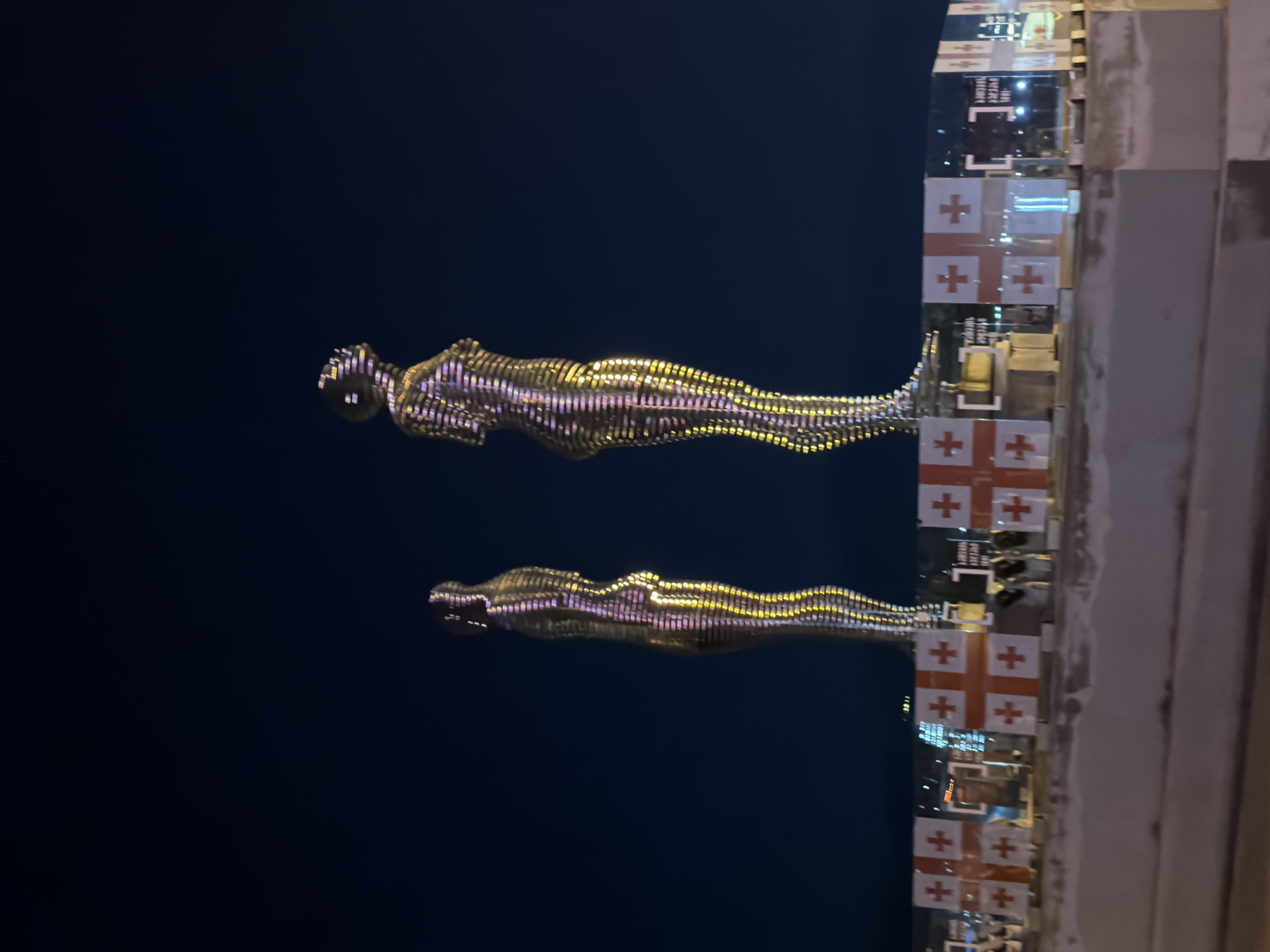 The image shows the Ali and Nino statue in Batumi, Georgia, at night. The two metallic figures are illuminated and appear to be rotating, silhouetted against the dark sky. Adjacent to the figures is a structure decorated with multiple Georgian flags. The cityscape is visible in the background.