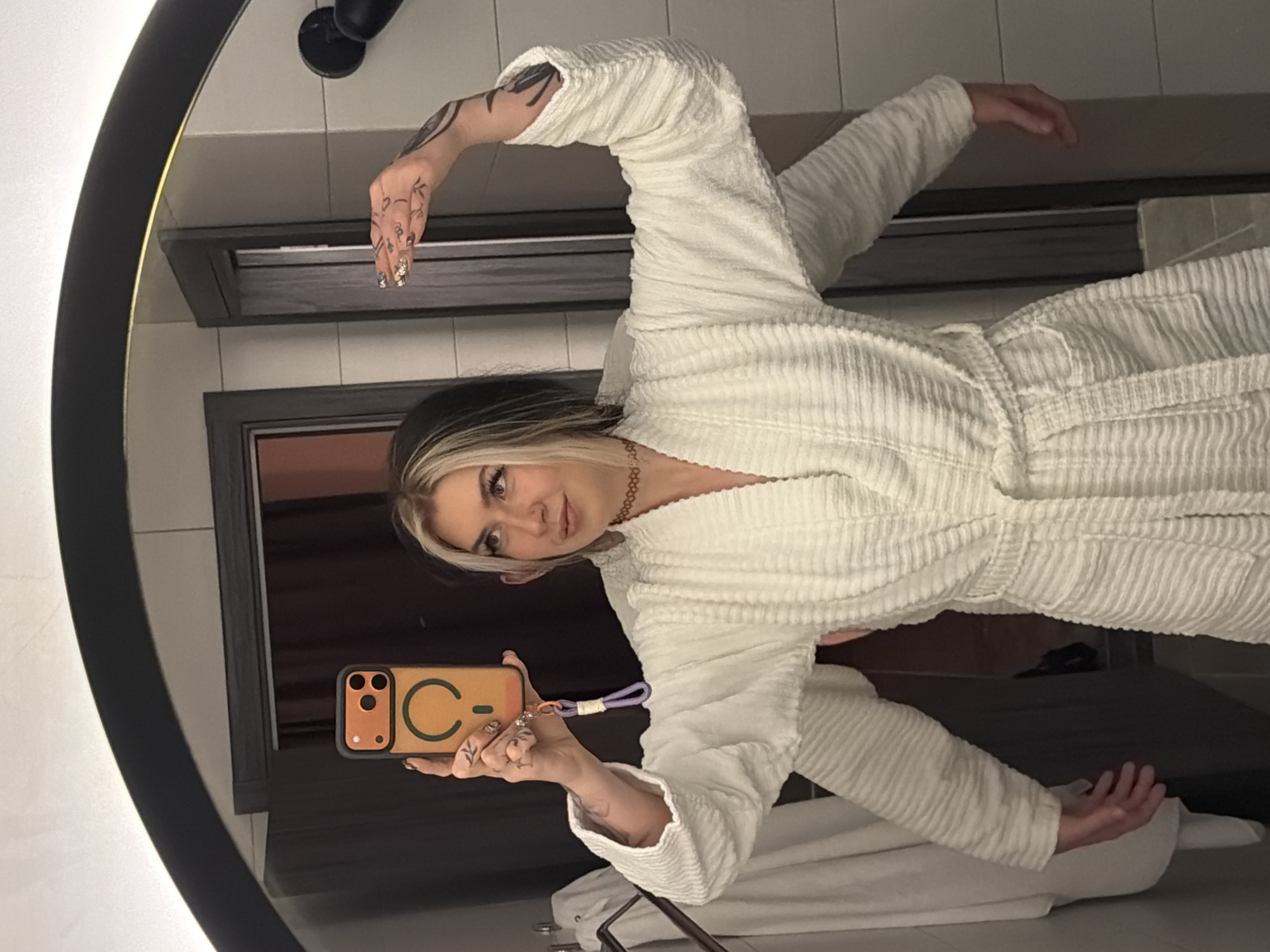 A woman wearing a white robe is taking a selfie in a mirror. The mirror is circular and framed in black. She is holding an orange iPhone in her left hand. She has blonde streaks in her hair. She is also wearing a necklace and has tattoos on her arms and hands. The background appears to be a hotel bathroom. The location cannot be determined from the image itself; the city of Palilula Urban Municipality, Serbia, cannot be confirmed or refuted.