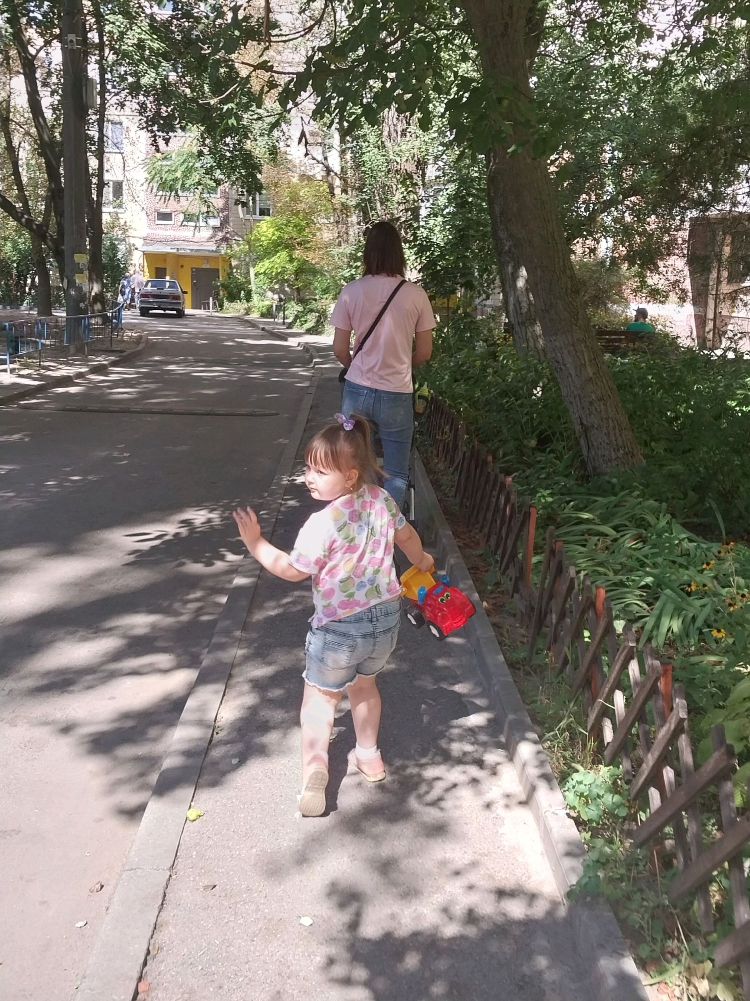 A young girl, wearing a pink and white fruit-patterned t-shirt and denim shorts, runs along a sidewalk. She is pulling a red and yellow toy truck. Her right arm is raised, and she appears to be looking over her shoulder. Behind her, an adult, seen from the back, walks on the same sidewalk. The adult has long brown hair and wears a pink t-shirt and blue jeans. They are both walking away from the camera. The sidewalk runs alongside a paved road with a car visible in the distance. To the right of the sidewalk is a low wooden fence with a garden area behind it, featuring green plants and some yellow flowers. Trees with green foliage cast shadows across the scene. The background includes a multi-story building with a yellow section. The overall setting appears to be an urban residential area, possibly a courtyard or street in Dnipro, Ukraine, given the architectural style and vegetation.