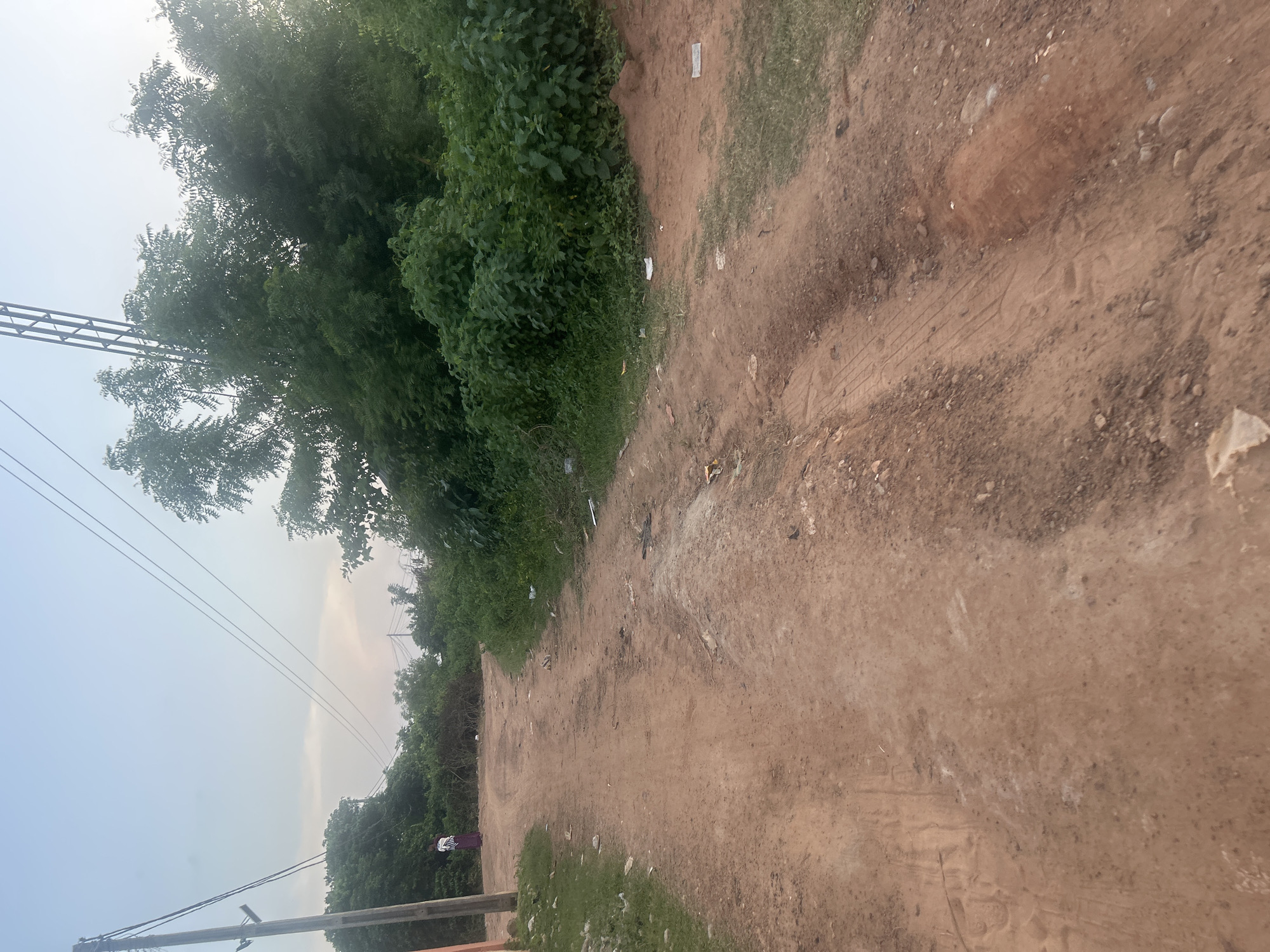 The image depicts a dusty, unpaved road winding through a rural or semi-rural area, likely in Ogbomosho, Nigeria. The road is marked with tire tracks and shows signs of wear and tear, with some unevenness and small rocks scattered across its surface. On the left side of the road, a utility pole stands tall, supporting power lines that stretch across the frame. A single individual, dressed in a patterned garment, is visible standing near the utility pole, facing away from the camera.
The road is bordered by dense green vegetation, including bushes and trees, which create a natural boundary. The sky above is overcast, with a soft, diffused light suggesting either early morning or late afternoon. Scattered on the roadside are various pieces of litter, including white paper or plastic fragments, indicating a lack of consistent waste management. The overall scene conveys a sense of a typical, undeveloped road in a Nigerian setting, possibly a residential area or a path leading to agricultural land.