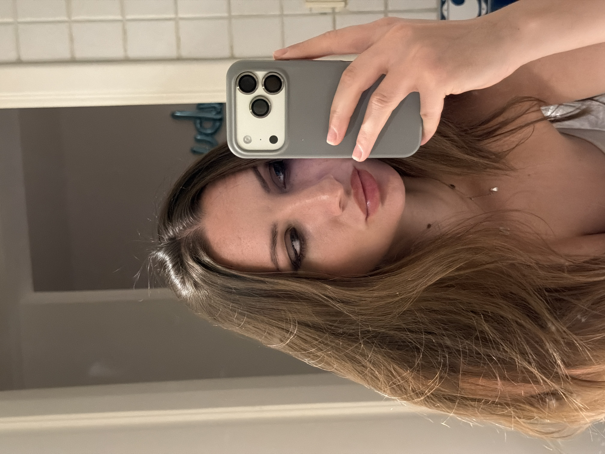 A woman is taking a selfie in a bathroom. She is holding a grey phone in her right hand, which is reflected in the mirror. She has long, wavy brown hair and is wearing makeup. Her gaze is directed towards the phone. The bathroom appears to have tiled walls and a light-colored doorframe. There is a blue decorative item with letters visible on the wall behind her. The image does not provide any information or context that would place it in Sharm El Sheikh, Egypt.