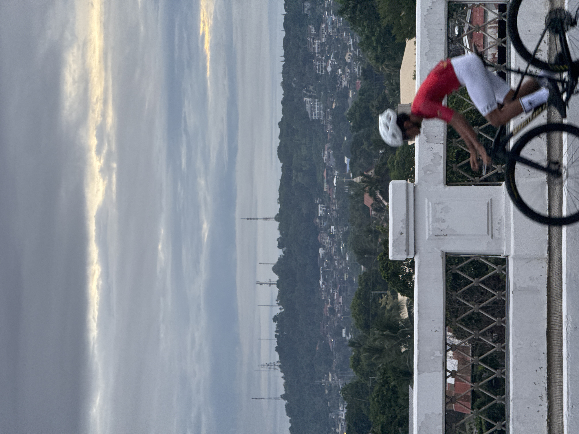 A medium shot, taken during overcast lighting conditions in Davao City, Philippines, showcases a cyclist positioned on a white painted concrete railing. The cyclist, wearing a white helmet, red shirt, and white shorts, is bent over their road bicycle. The railing features square windows with a diamond-shaped metal grid. In the background, there's a view of a hillside with buildings, trees, and tall telecommunications towers. The sky above is filled with grey clouds interspersed with hints of sunlight.