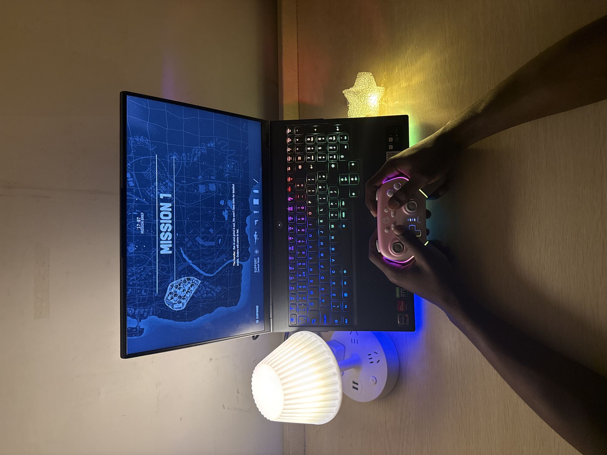 A dimly lit desk features a gaming laptop displaying "MISSION 1," with "Jiangjin, China" faintly visible on the screen. A gamer's hands hold a pink and neon-lit controller. A small mushroom-shaped lamp illuminates the scene from the lower-left corner. A star-shaped light is positioned behind the laptop. The backdrop consists of a plain wall and a wooden surface.