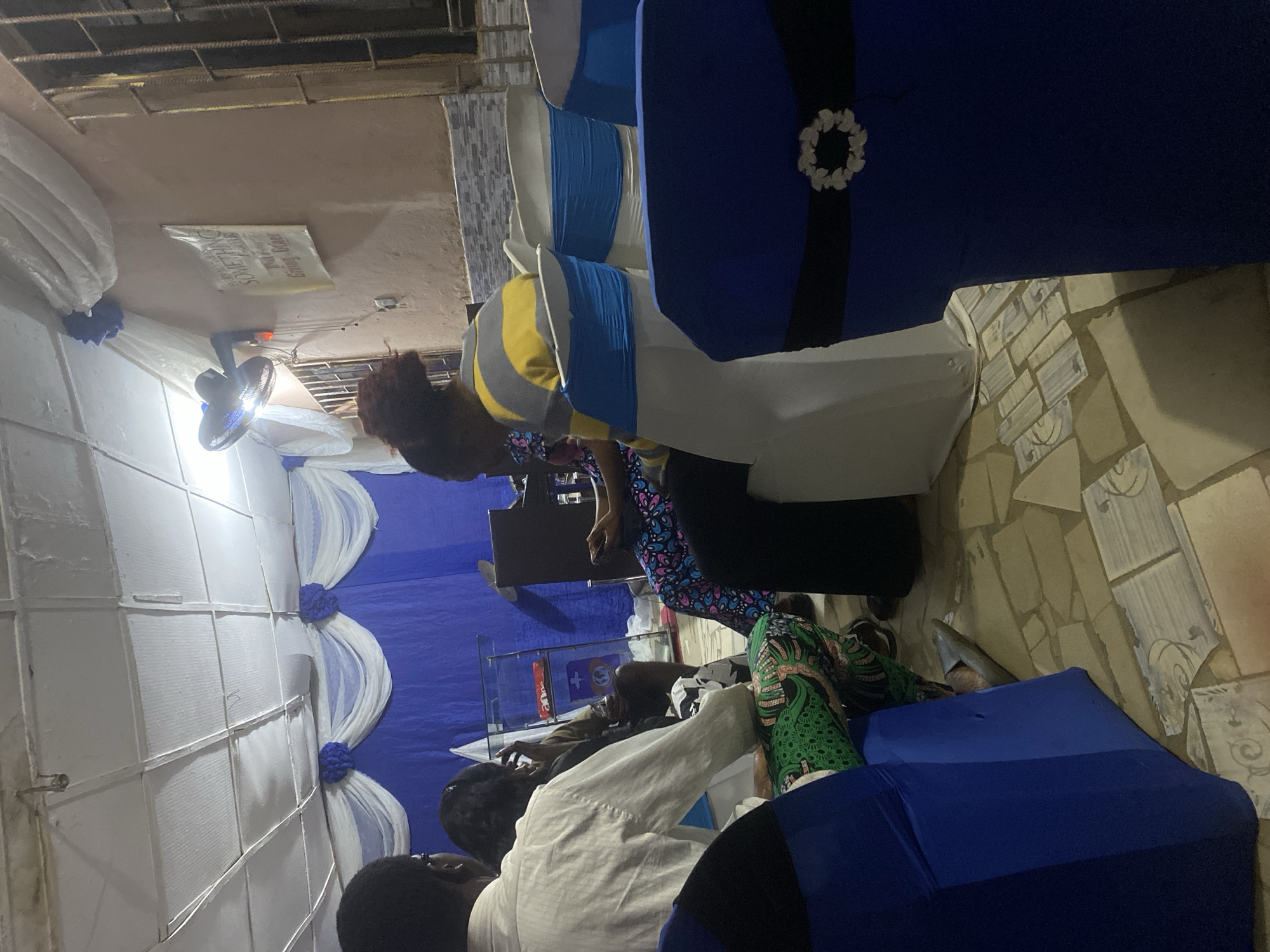 This image captures an indoor gathering in Bwari, Nigeria, characterized by a blue and white decorative theme. The setting features a white suspended ceiling with visible grid lines and a hook fixture in the upper left. The right wall is light brown with a textured finish, bordered by smaller patterned tiles below, and includes a window secured with vertical metal bars. A black oscillating fan with an integrated light is mounted on this wall, below which hangs a light-colored banner partially legible as "SOMETHING FOR STRATEGIC GIVING-OFFERING." The back wall is draped with blue fabric and adorned with white swags featuring blue floral clusters. The floor is tiled with irregular, light-toned tiles displaying subtle decorative patterns.
Approximately six individuals are present, seated in chairs. In the immediate foreground, a dark blue chair cover with a black sash and a white, floral-like buckle is prominent on the right. To the left, the back of another blue chair cover is visible. In the mid-ground, a person wearing a white long-sleeved shirt is seated on the left, beside another individual wearing vibrant green patterned trousers. On the right, a person with reddish-brown hair, dressed in a yellow, grey, and white striped shirt, is observed holding and interacting with a smartphone. Further back, near a clear acrylic pulpit with a red item on its lower shelf and a circular logo, two additional individuals are partially visible, one of whom is also engaging with a smartphone. The individuals are seated in chairs, some of which are covered in white fabric with blue sashes. The overall scene suggests a community event or service.