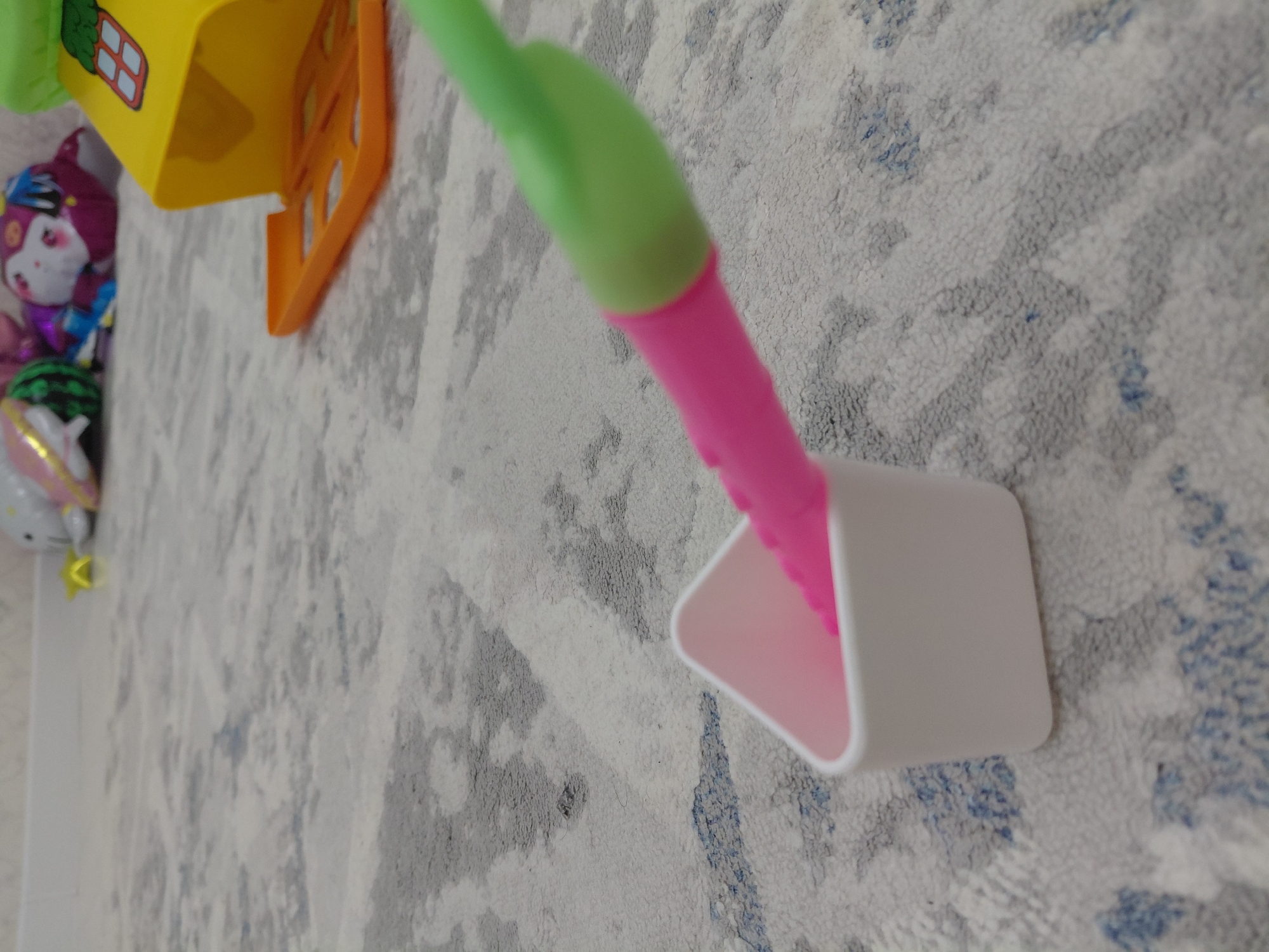 An overhead shot shows a white triangular-shaped toy, with a pink and green toy stick partially inside it, lying on a light gray carpet with a subtle blue and dark gray pattern. In the background, a yellow toy house with an orange door is visible, along with out-of-focus, colorful balloons. The scene appears to be indoors, in a domestic setting, with no indication of the city of Aktau, Kazakhstan.