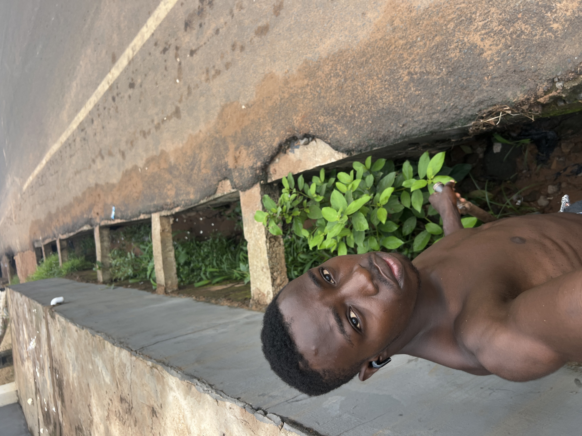 A shirtless young Black man with short, dark hair and a mustache is standing next to a concrete drainage ditch on a paved road. He is looking at the camera and wearing a black earbud in his left ear. The drainage ditch is filled with green foliage and some litter. The road has a white line painted down the center and appears to be in an urban or semi-urban area. The man is holding a light-colored object in his right hand. This scene is located on a street in Nkpor, Nigeria.